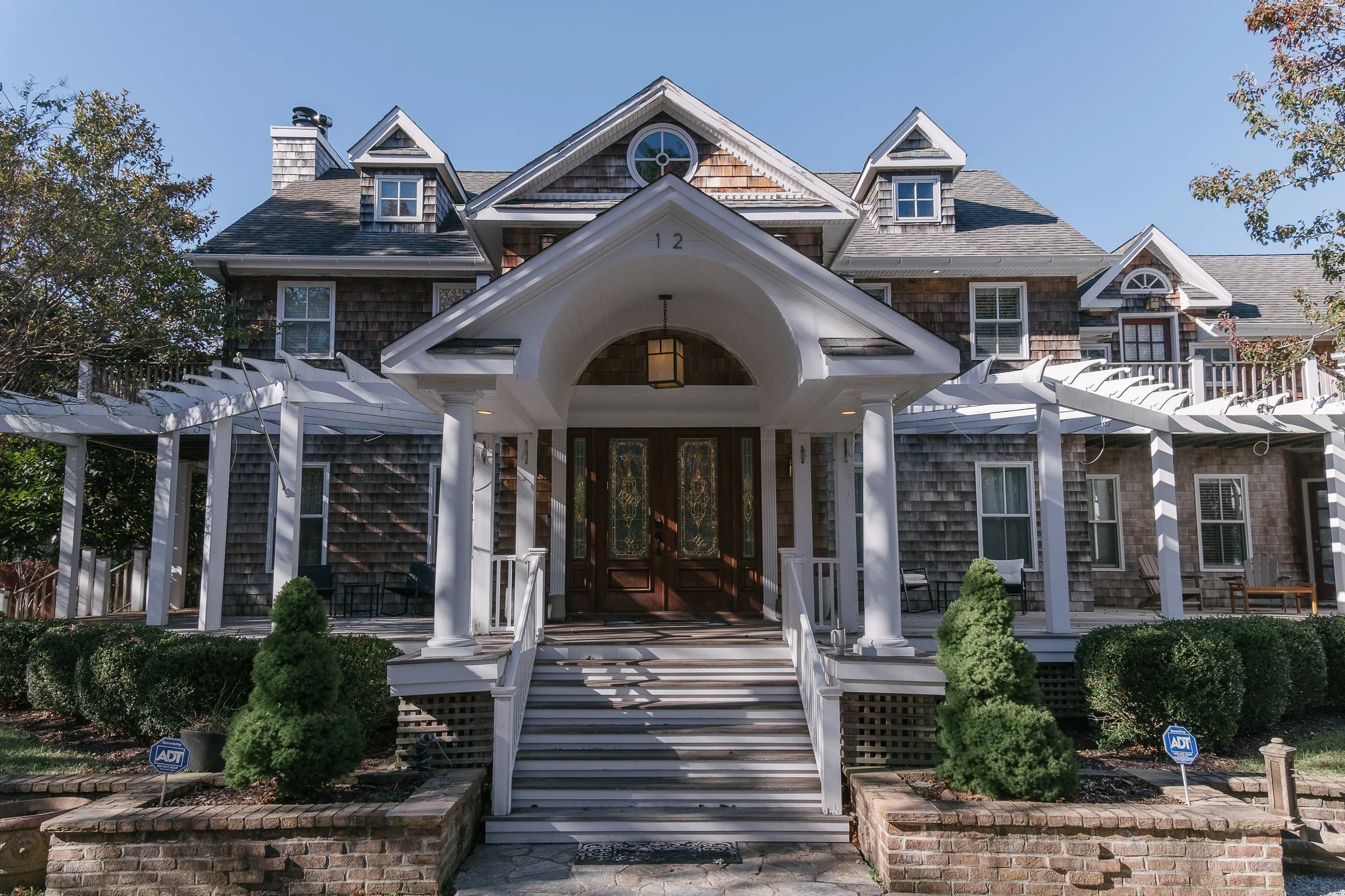 Large multi-story house with steep gabled roof, wooden shingles, and a covered front porch with stairs and white columns, surrounded by green bushes and trees.