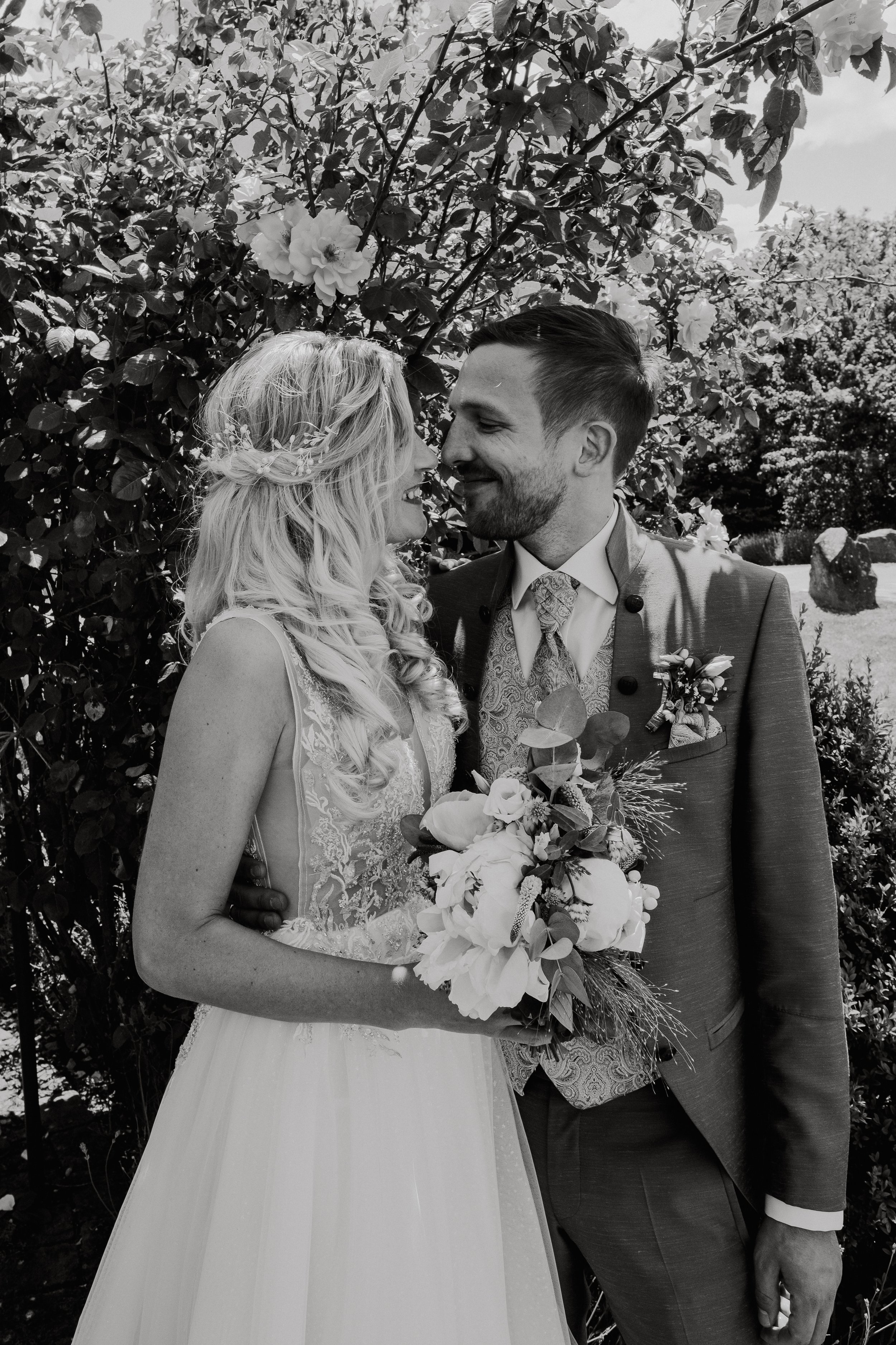A black and white photo of a wedding couple standing close together outdoors, smiling, with a background of bushes and trees. The bride is holding a bouquet and has long, wavy blonde hair, while the groom is dressed in a suit with a patterned tie and