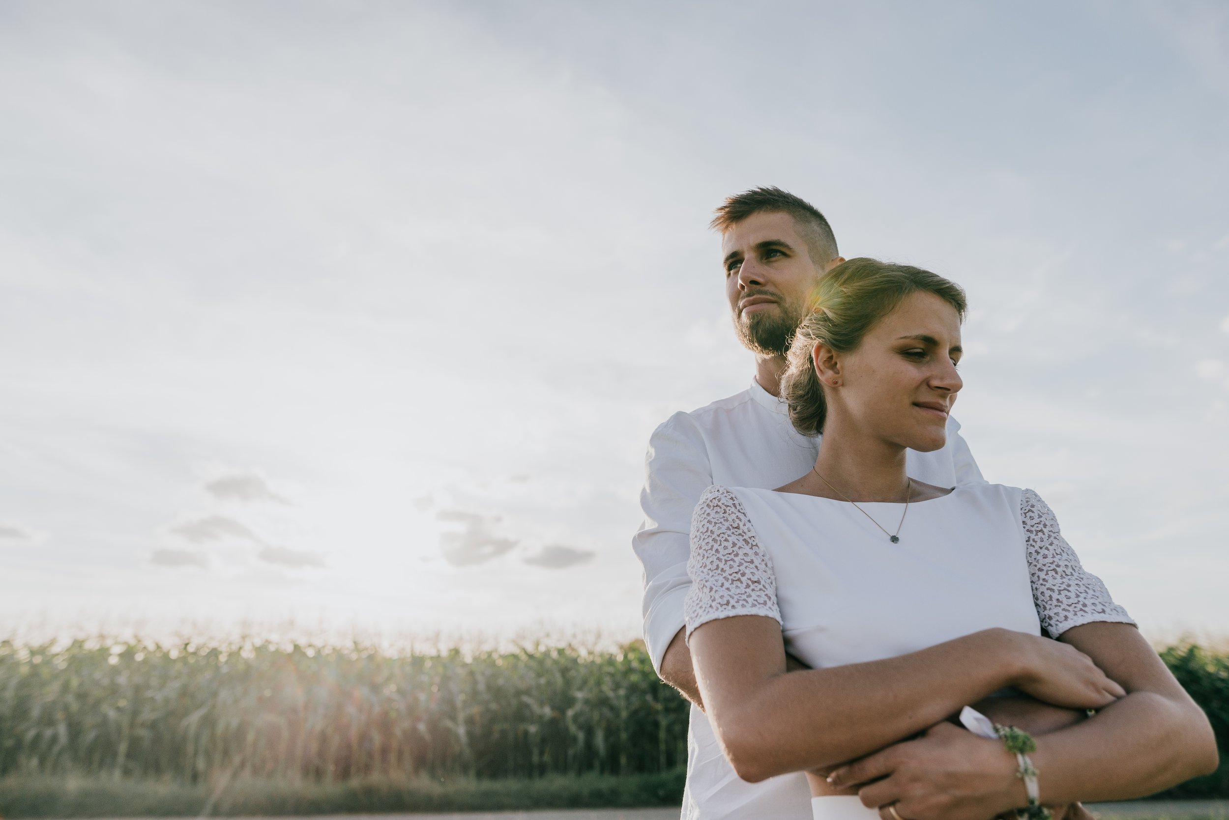 A young couple stands outdoors during sunset, with the man embracing the woman from behind. The woman has her arms crossed and is wearing a white dress with lace sleeves, and the man is dressed in a white shirt. The sky is partly cloudy, and a field