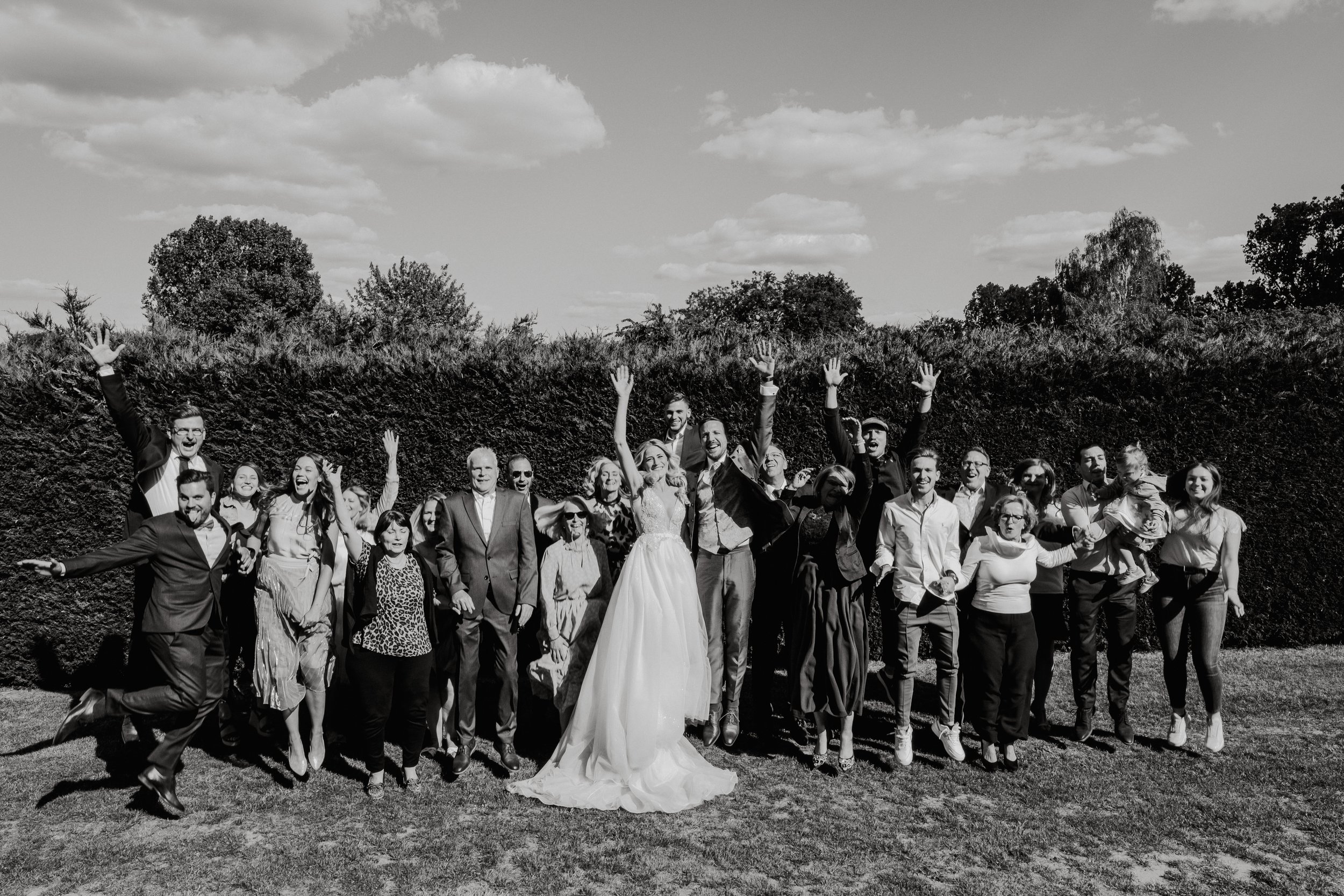 A group of people celebrating outdoors in a sunny setting at their destination wedding, with some raising their hands and smiling, including a bride and groom in the center, all dressed in formal attire.