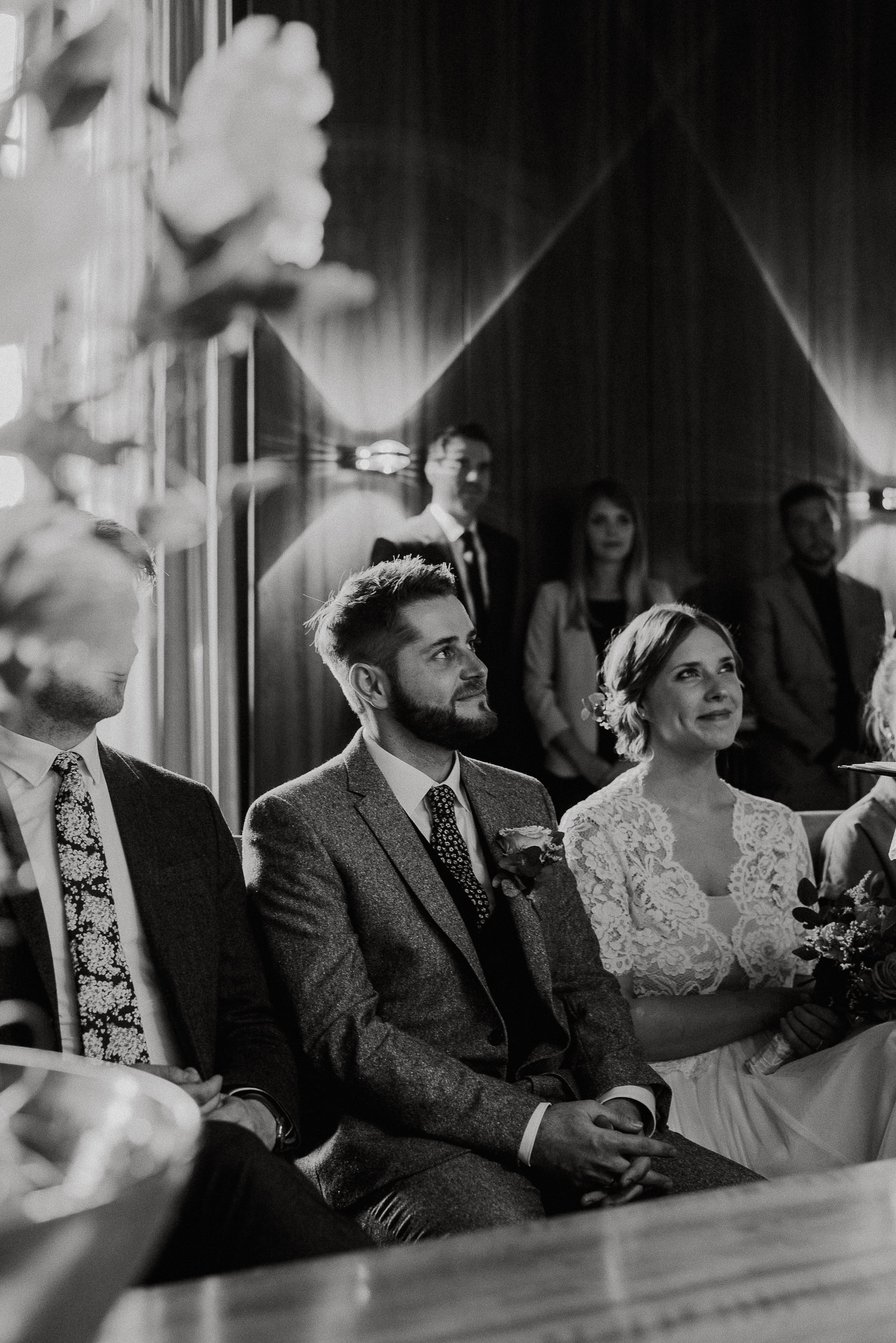 Black and white photo of a wedding ceremony with the bride and groom sitting in front. The groom has a beard and is wearing a suit, while the bride is in a lace wedding dress holding a bouquet. Other guests are visible in the background, standing and