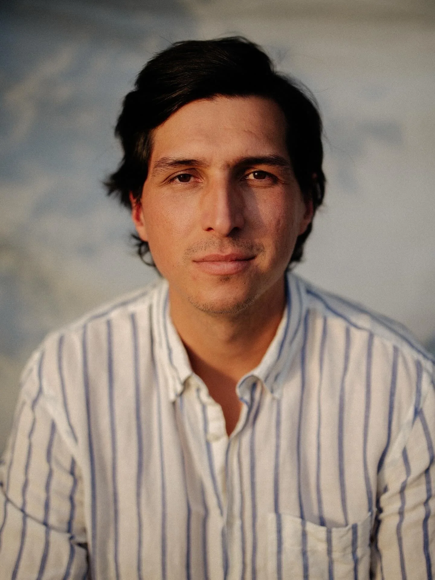 Jair Rojas, the founder of Amauri May Studio wearing a white and blue striped shirt, looking at the camera with a neutral expression, outdoors during sunset or sunrise in a Hamptons portrait shooting.