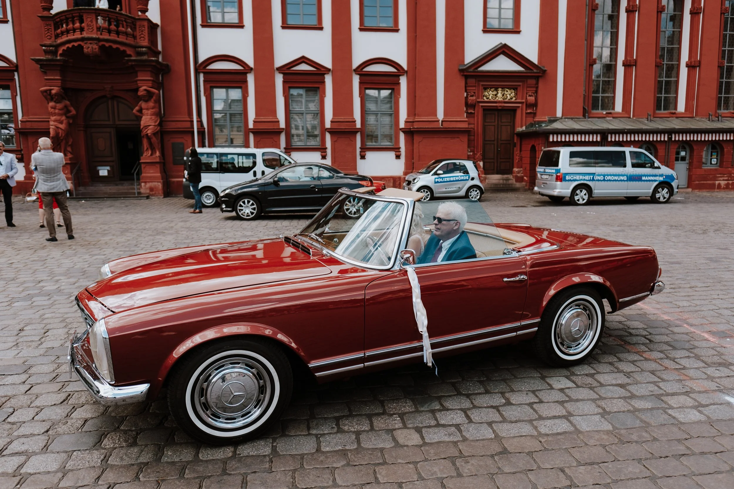 A vintage red convertible Mercedes-Benz car parked on a cobblestone street in front of a historic red brick building. A man with sunglasses and gray hair is sitting in the driver's seat of the car. Several people are standing and talking nearby, and