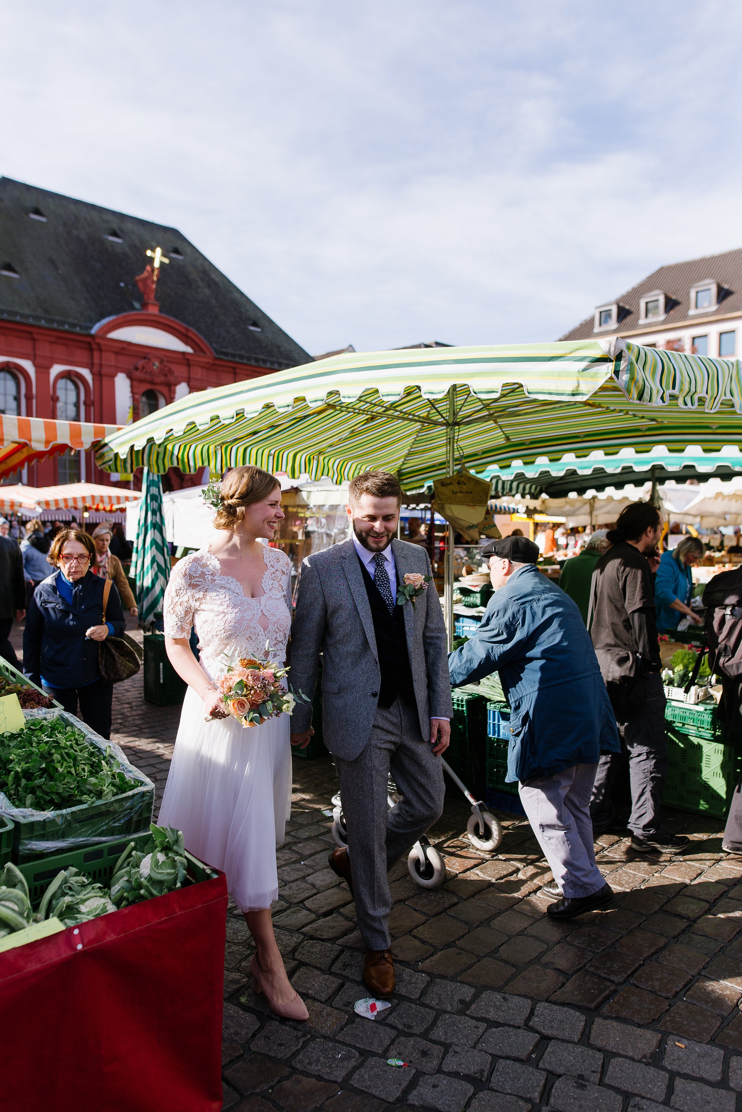 A bride and groom shopping at an outdoor market, holding hands and smiling, with shoppers and market stalls around them.