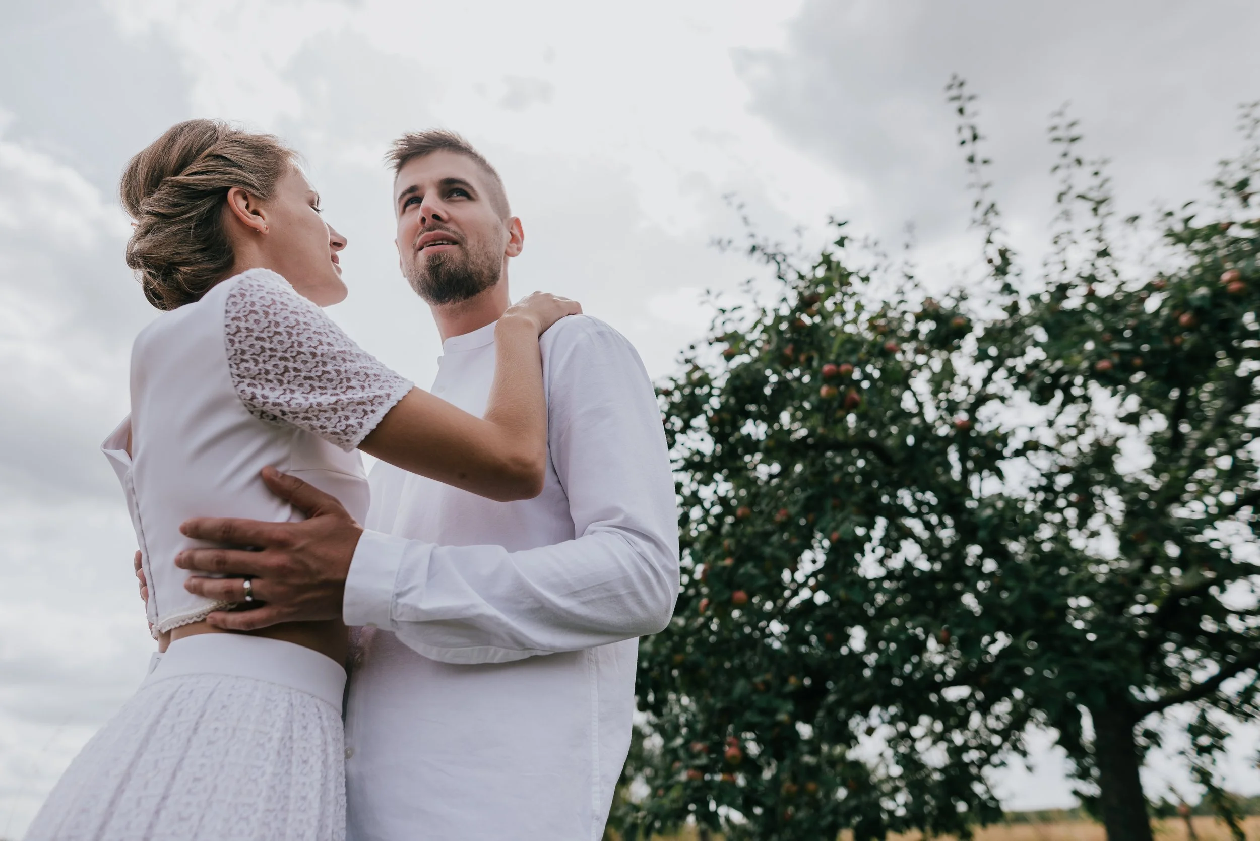 A couple embracing outdoors near an apple tree with a cloudy sky in the background.