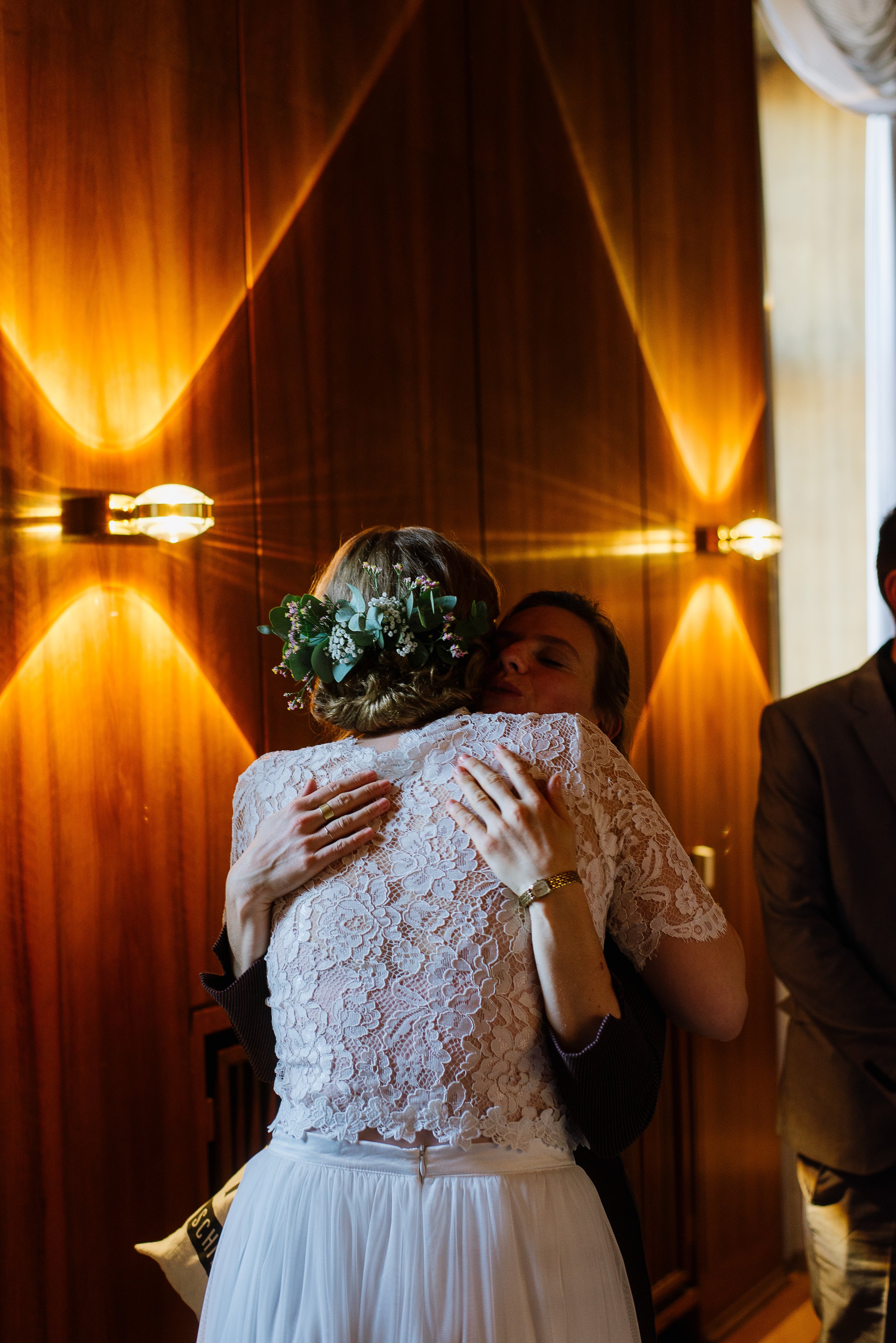 Two women hugging at a wedding celebration, one wearing a lace dress and a floral headpiece, the other in dark clothing, in a warmly lit room with wood-paneled walls.