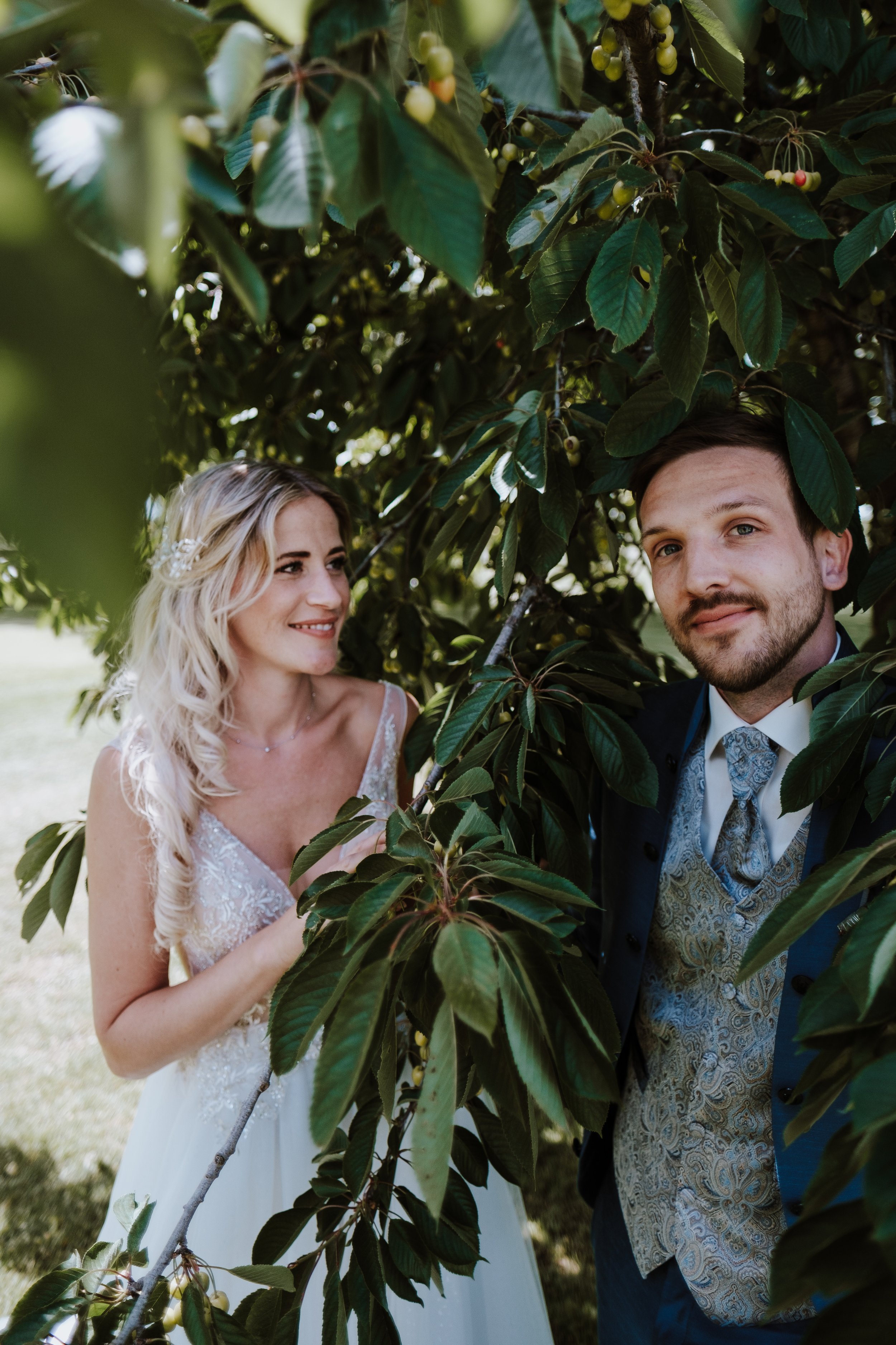 A bride and groom peeking from behind a tree with green leaves, outdoors on a sunny day at their elopement shoot.