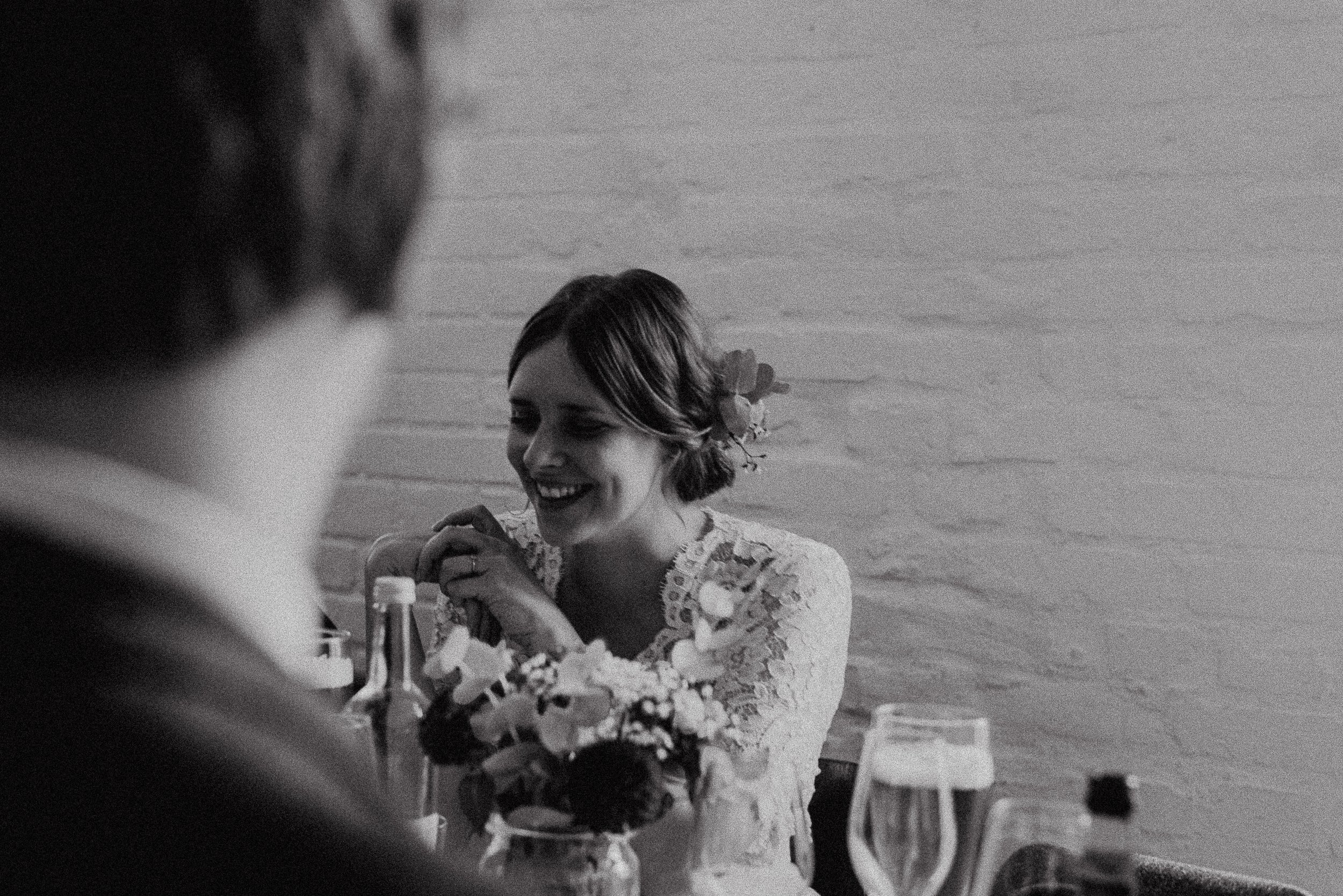 A woman with short hair smiling and wearing a lace dress, sitting at a table with a bouquet of flowers and drinks, in a black and white photo.