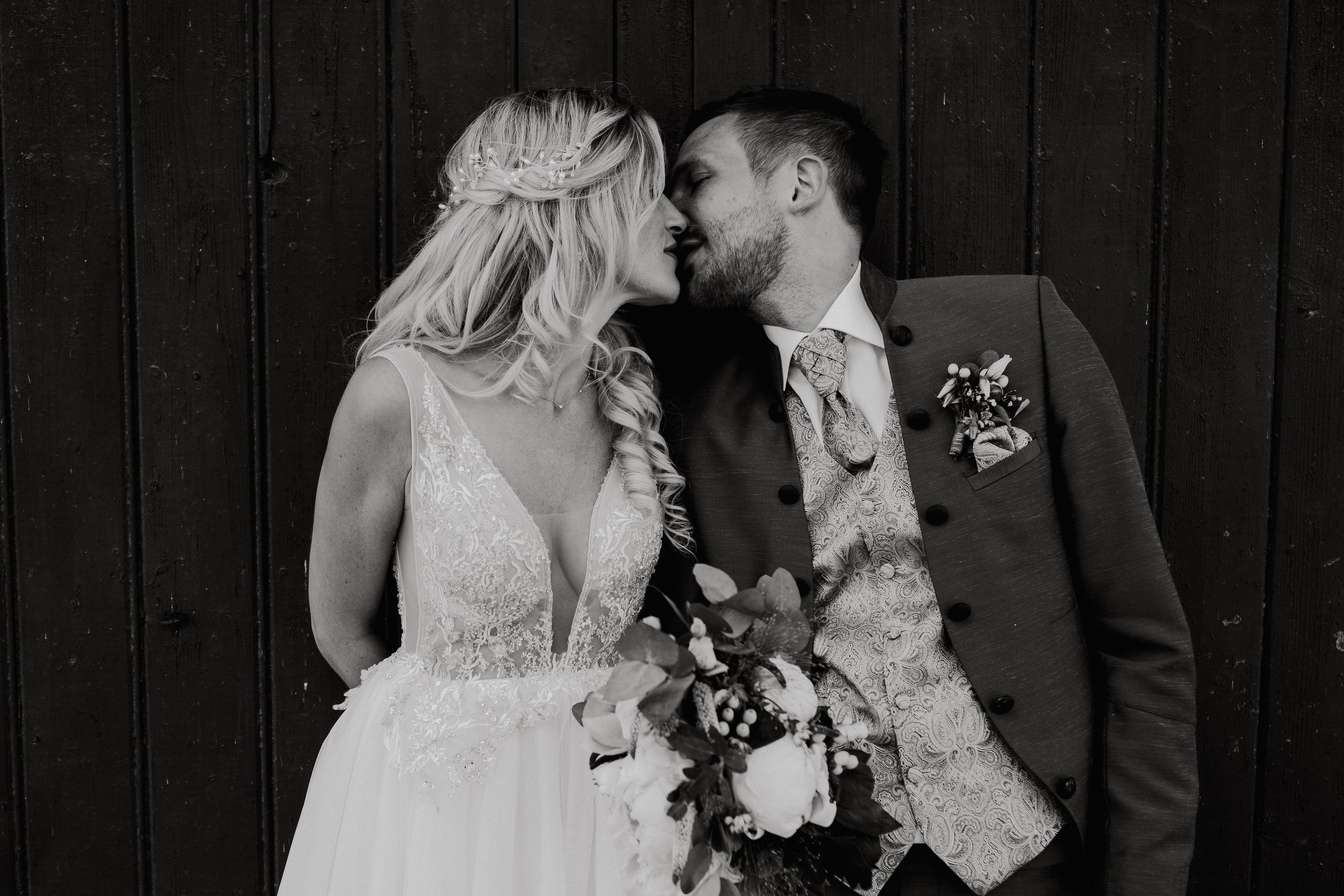 Black and white photograph of a bride and groom leaning in for a kiss, standing in front of a wooden fence. The bride wears a lace wedding dress with a plunging neckline, and the groom is dressed in a patterned vest and dark jacket with boutonniere.