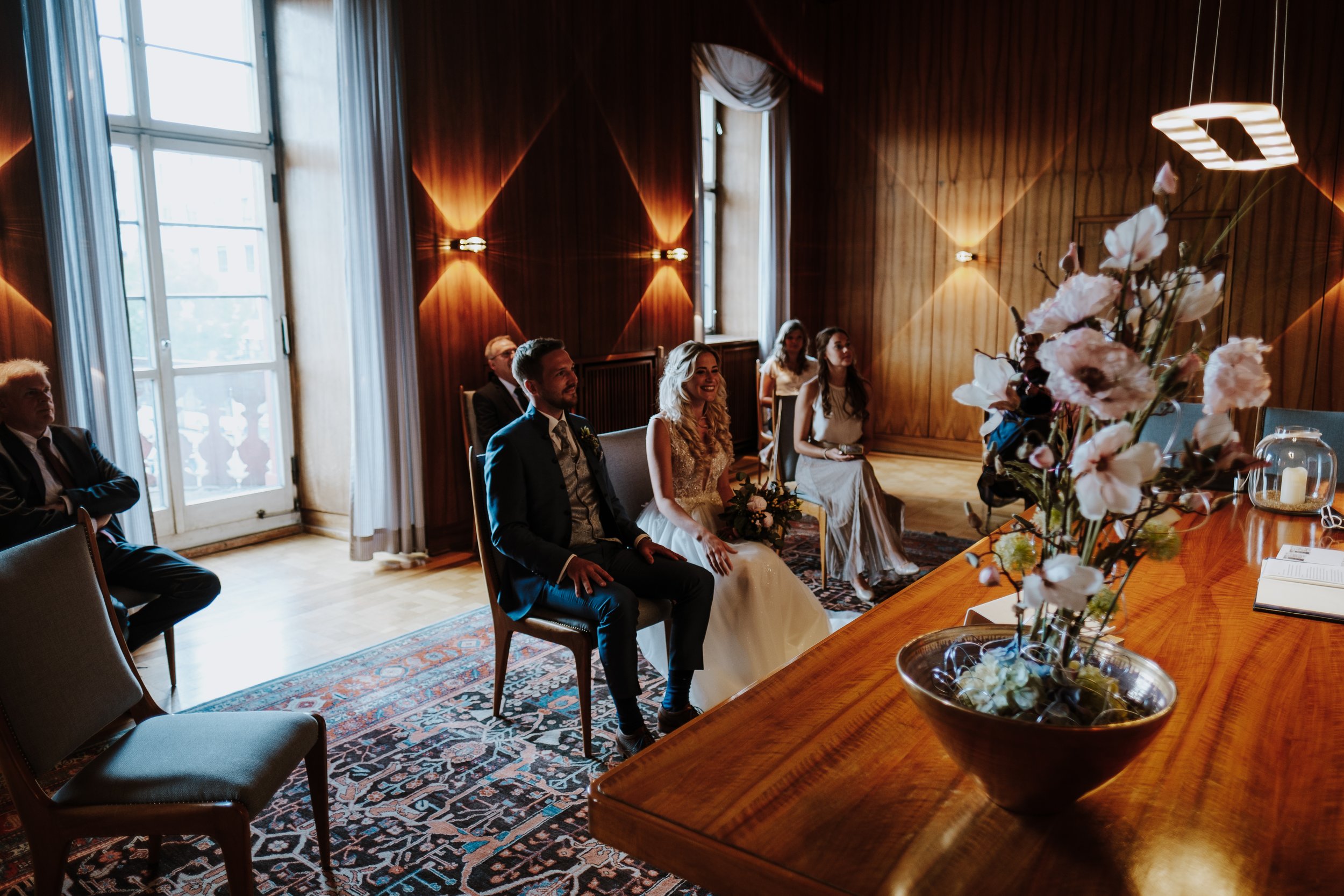 Wedding ceremony with bride, groom, and guests seated in a warmly lit wood-paneled room, with large windows, floral arrangements, and a wooden table.
