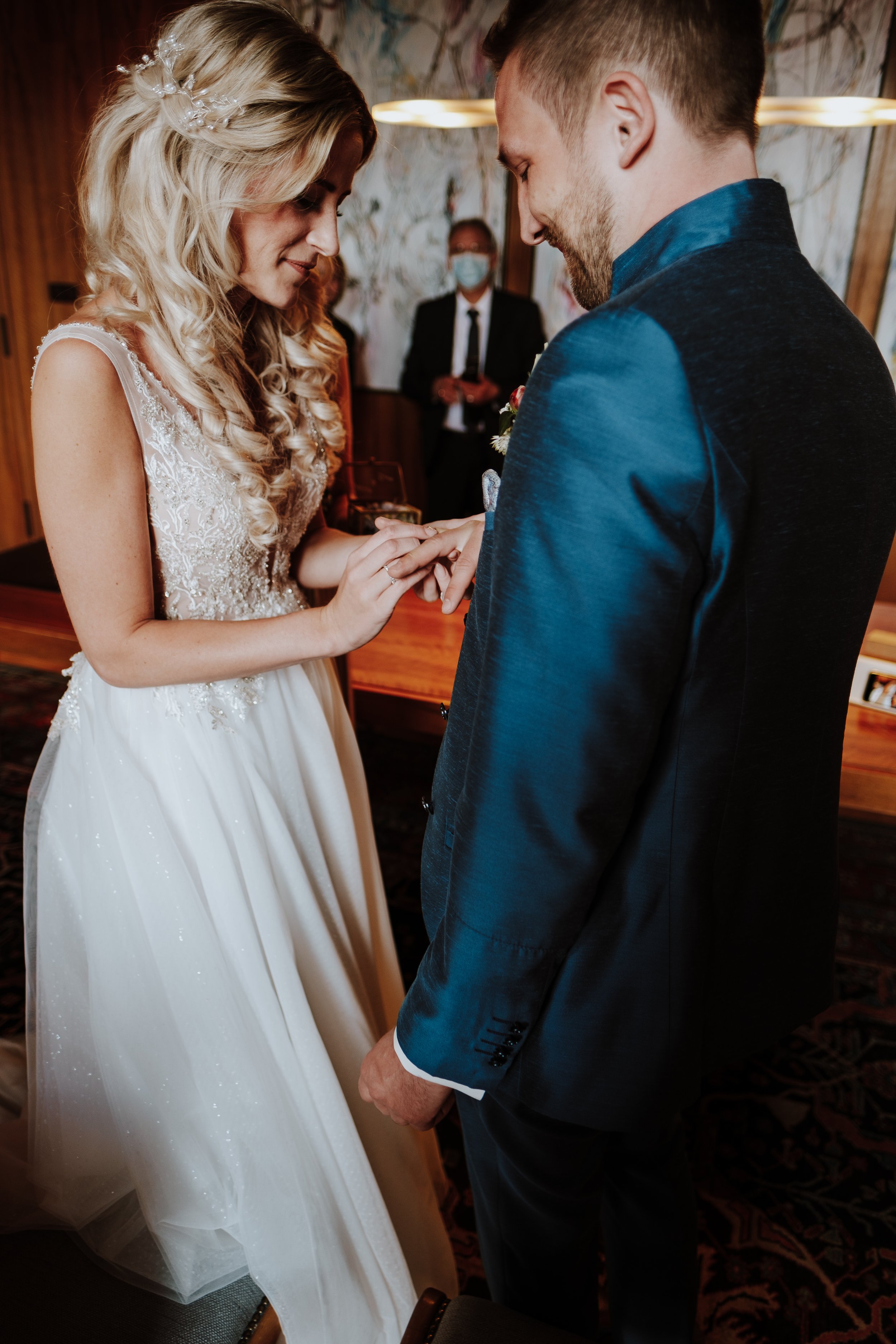 A woman in a white wedding dress is placing a wedding ring on a man's finger during a wedding ceremony, with a man in a suit in the background.
