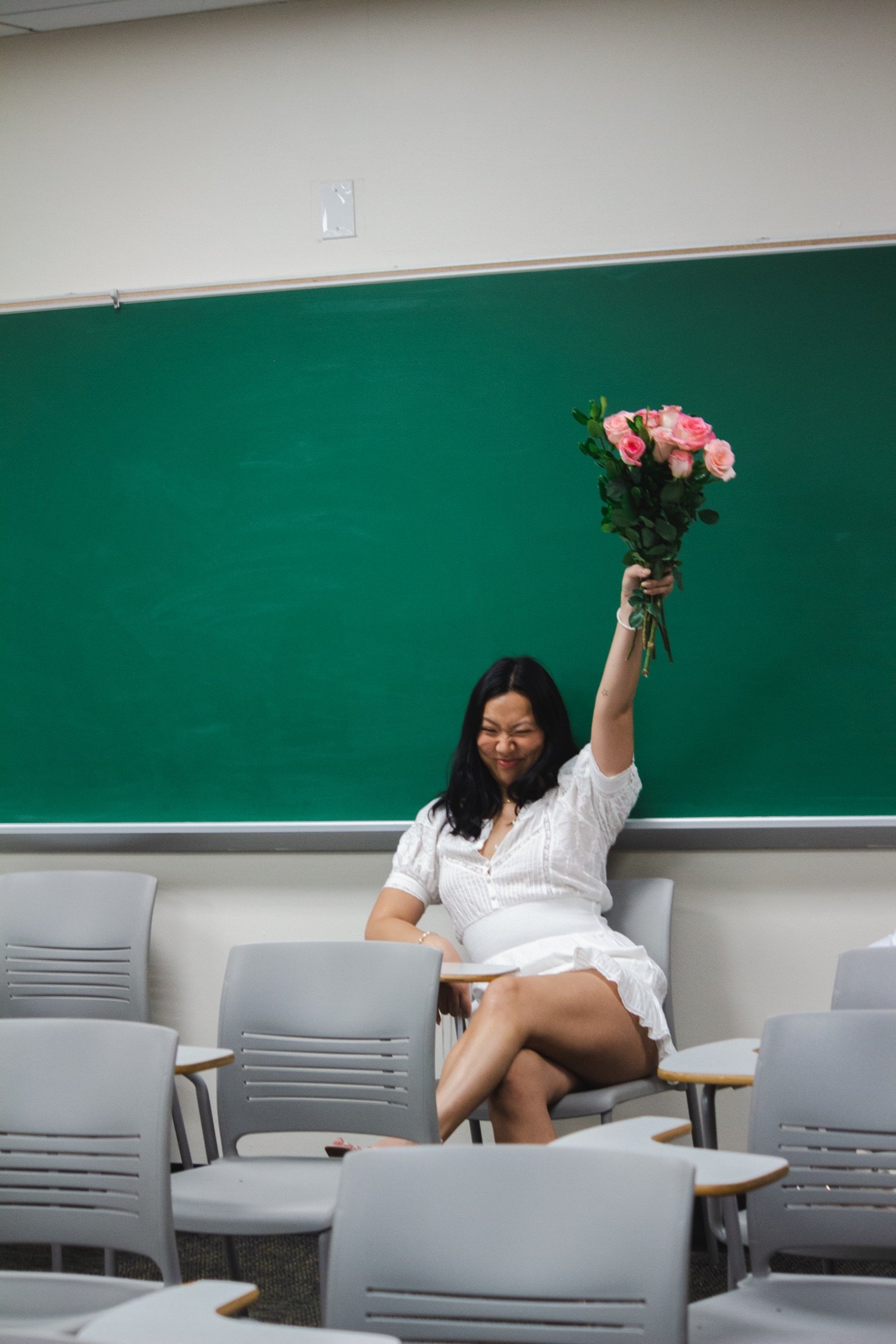 A woman sitting in a classroom, smiling with eyes closed, holding a bouquet of pink roses above her head, wearing a white dress.