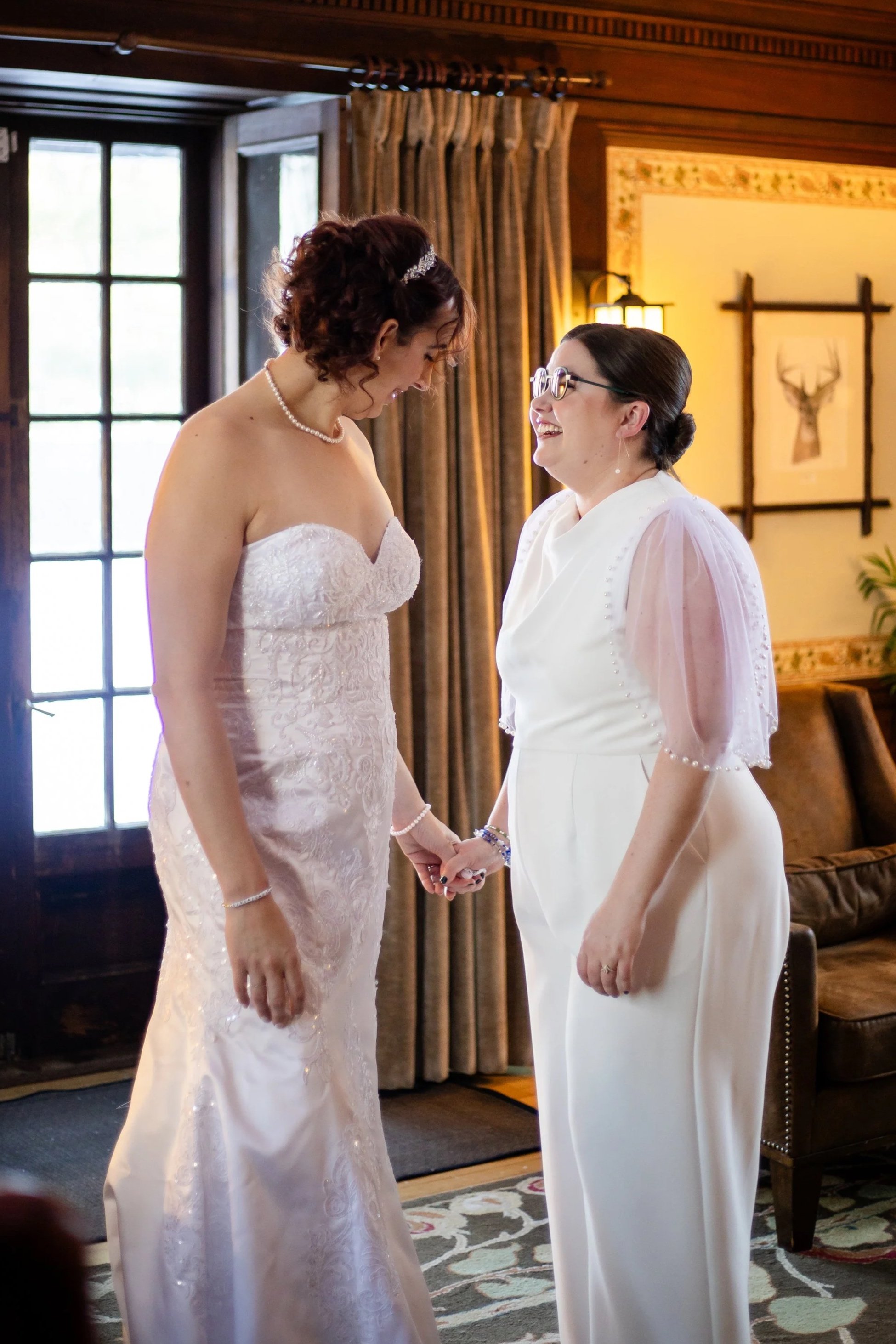 Two women in wedding dresses holding hands and smiling at each other indoors, with wooden paneling, curtains, and artwork in the background. Shot at the Willowdale Estate in Topsfield, Massachusetts, Greater Boston Area.