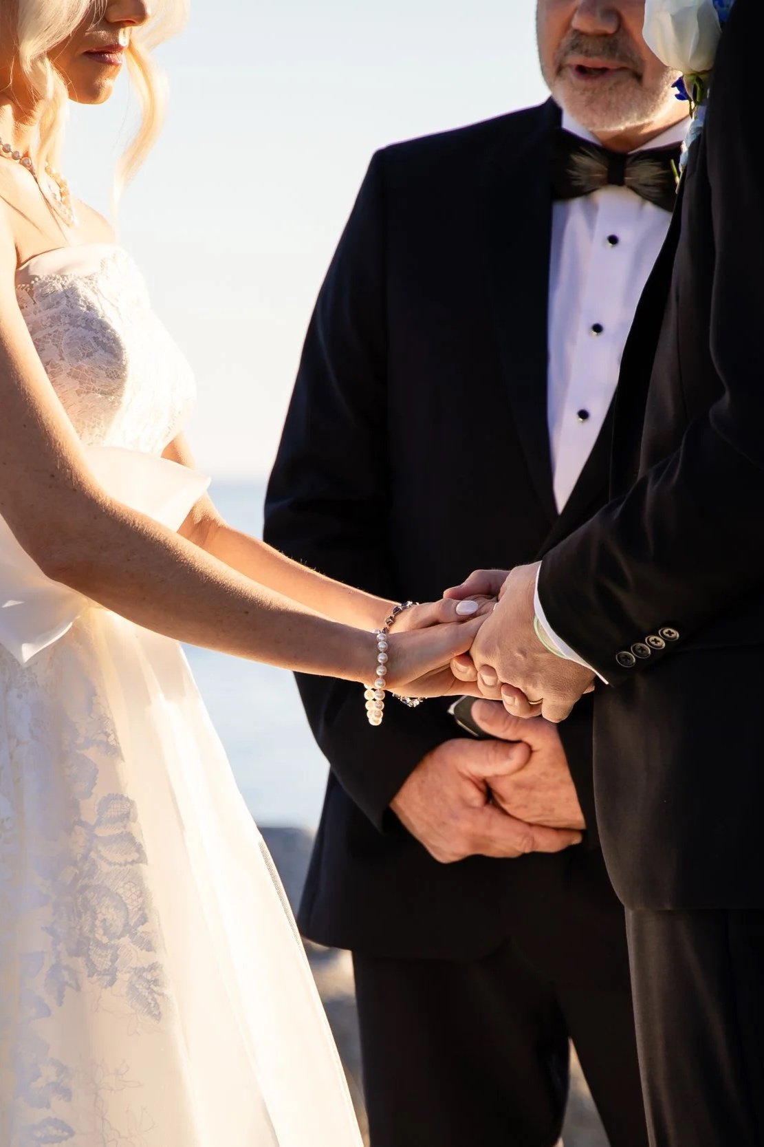 A bride and groom holding hands during their wedding ceremony, with an officiant standing behind them. The bride wears a lace wedding dress and pearl jewelry, and the groom is in a black tuxedo. Shot on the water on Seabrook Island, South Carolina.