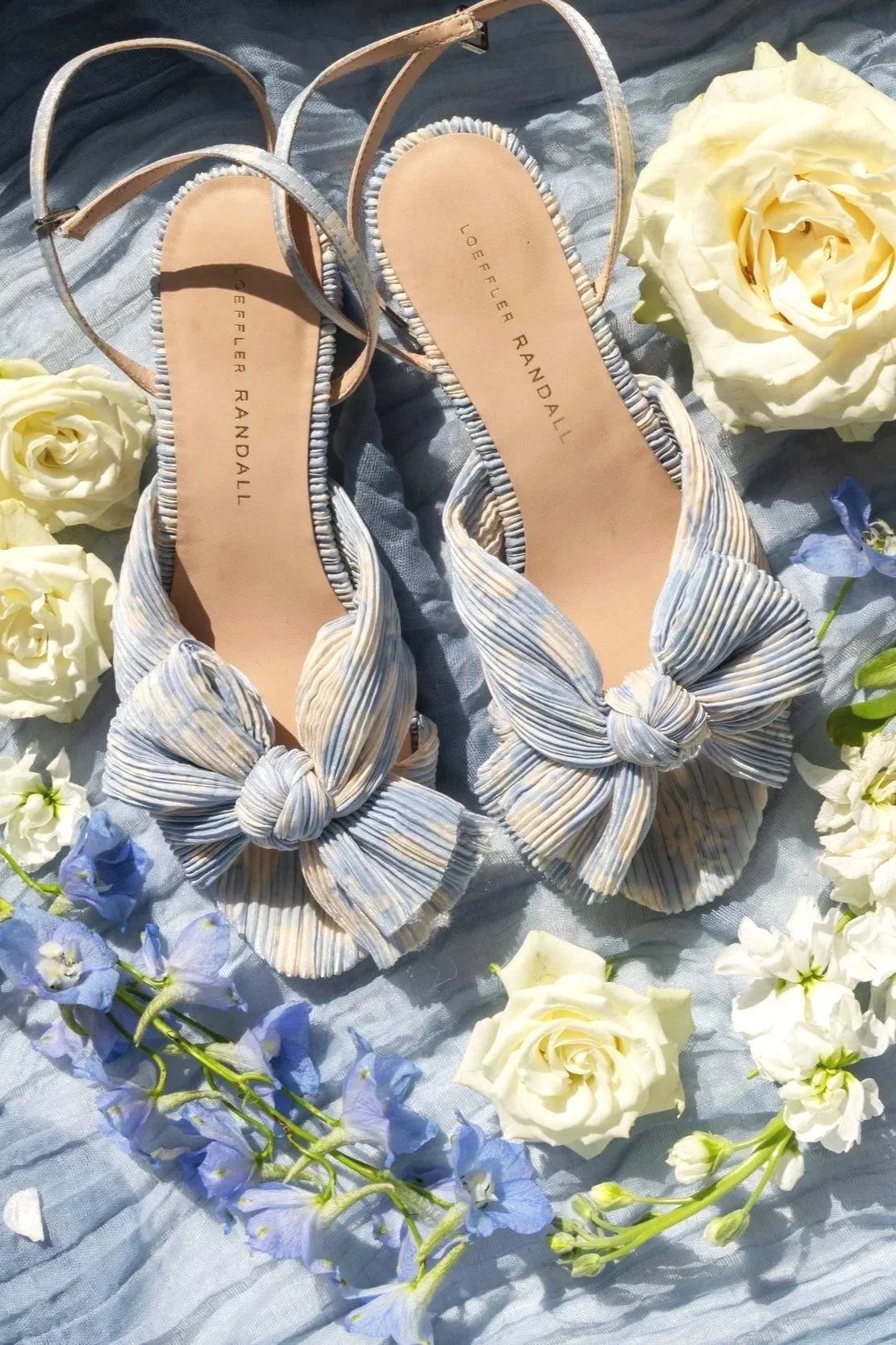 A pair of stylish striped women's shoes with large bow accents, displayed on a light blue fabric surrounded by white and light purple flowers.
