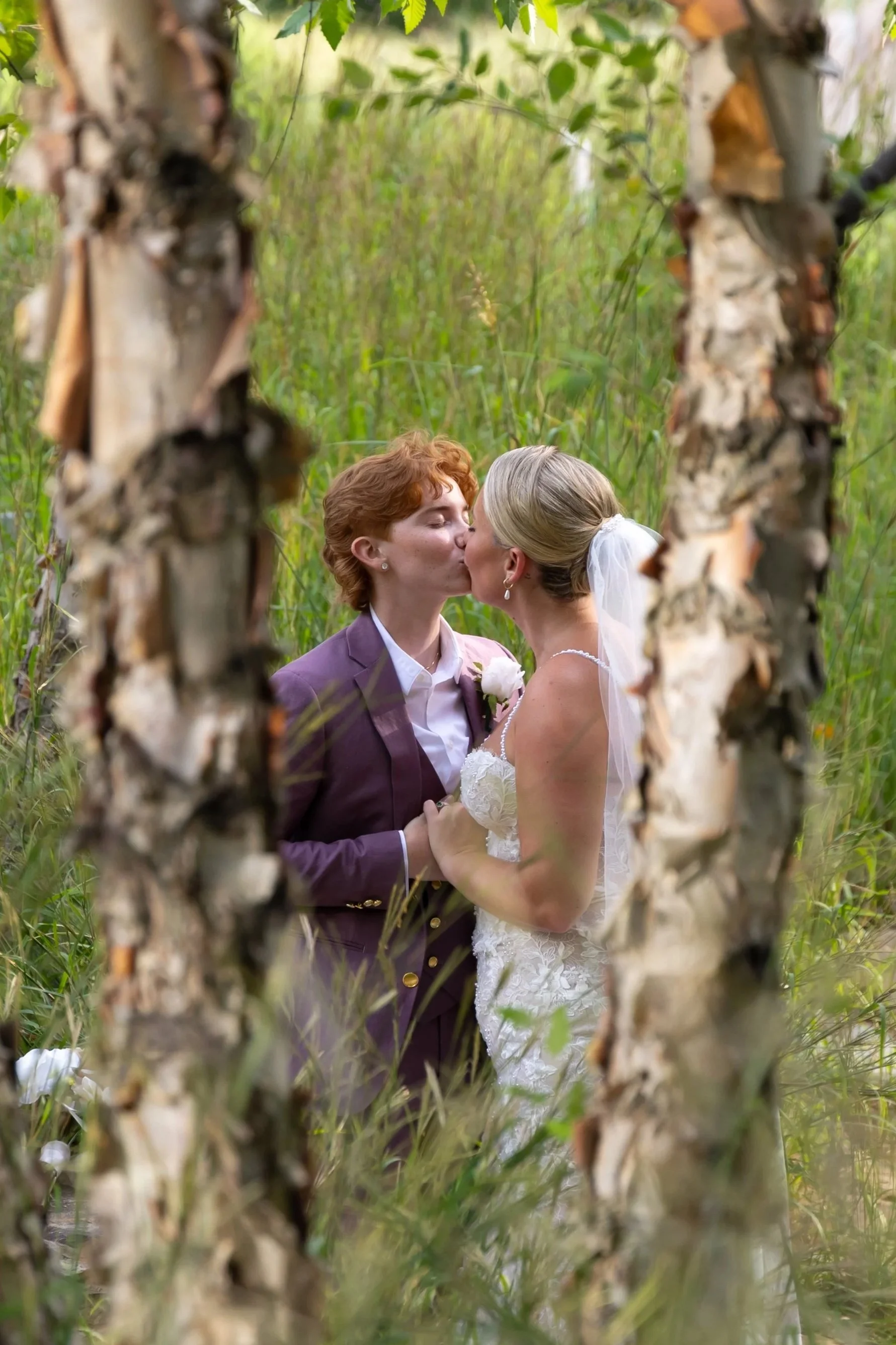 Two women sharing a kiss in a natural outdoor setting. The photo is taken through a frame of tree trunks and green foliage, creating a natural border around the couple. Shot at The Row Hotel at Assembly Row in Somerville, Massachusetts.