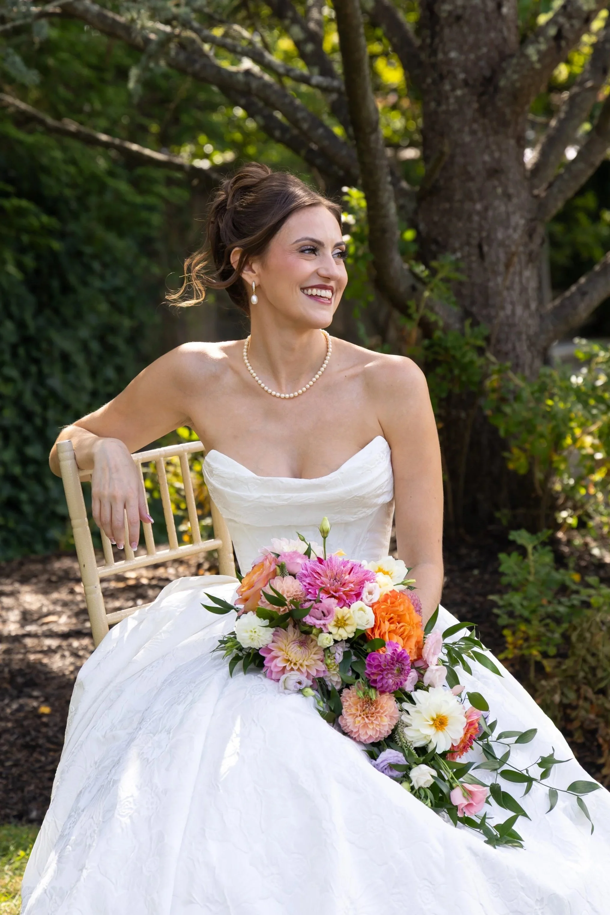 A smiling woman in a white wedding dress sitting outdoors near a tree, holding a colorful bouquet of flowers.