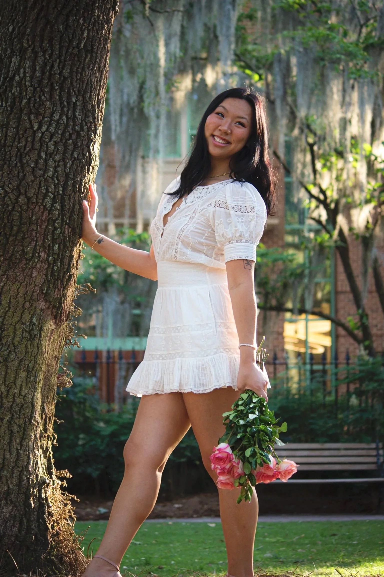 A woman in a white dress holding pink roses standing outdoors near a tree in a park with trees and a bench in the background.