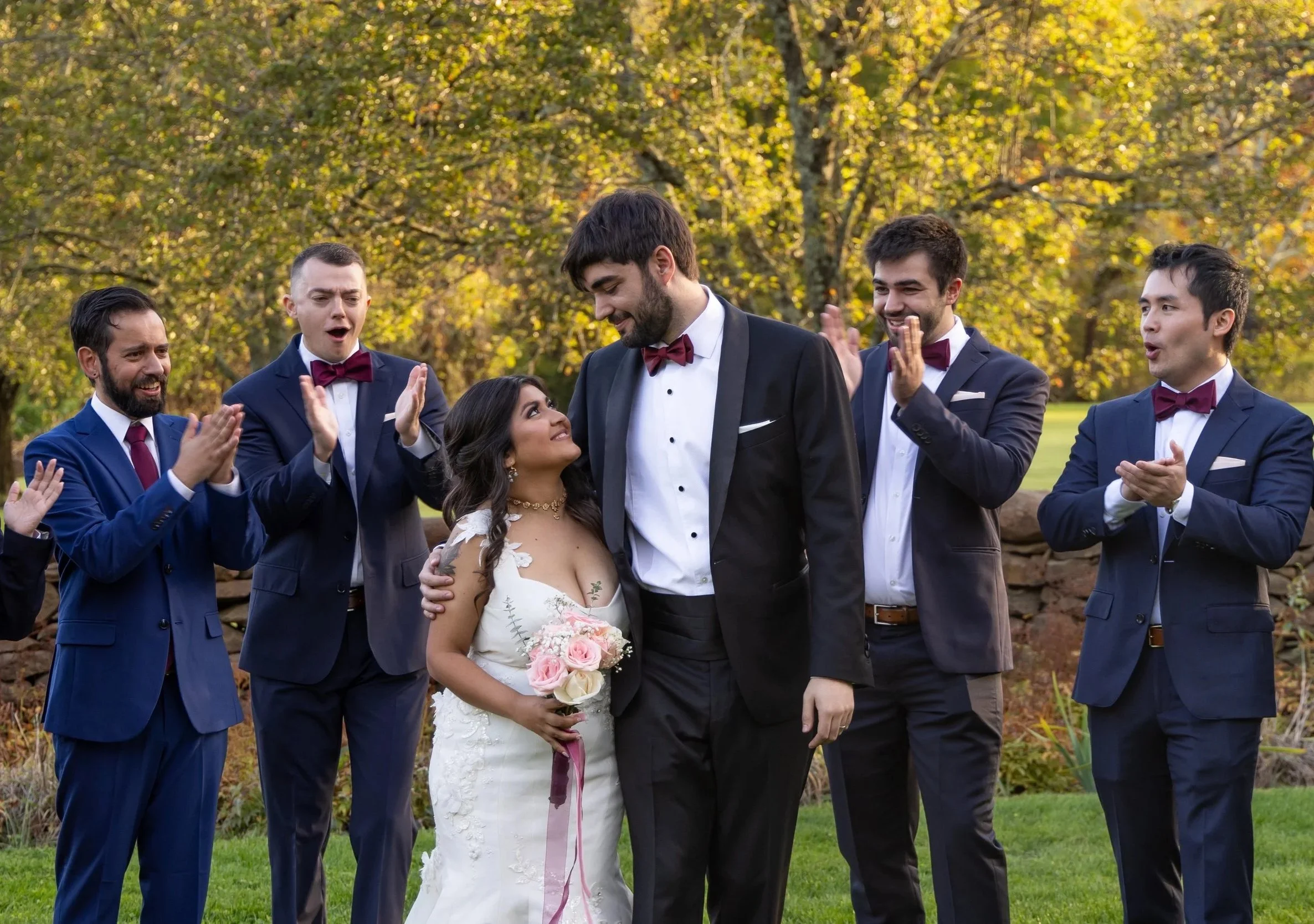 A bride and groom standing outdoors surrounded by five groomsmen applauding and looking at them, with trees and yellow foliage in the background at Independence Harbor owned by Sapphire Event group in Assonet, MA.