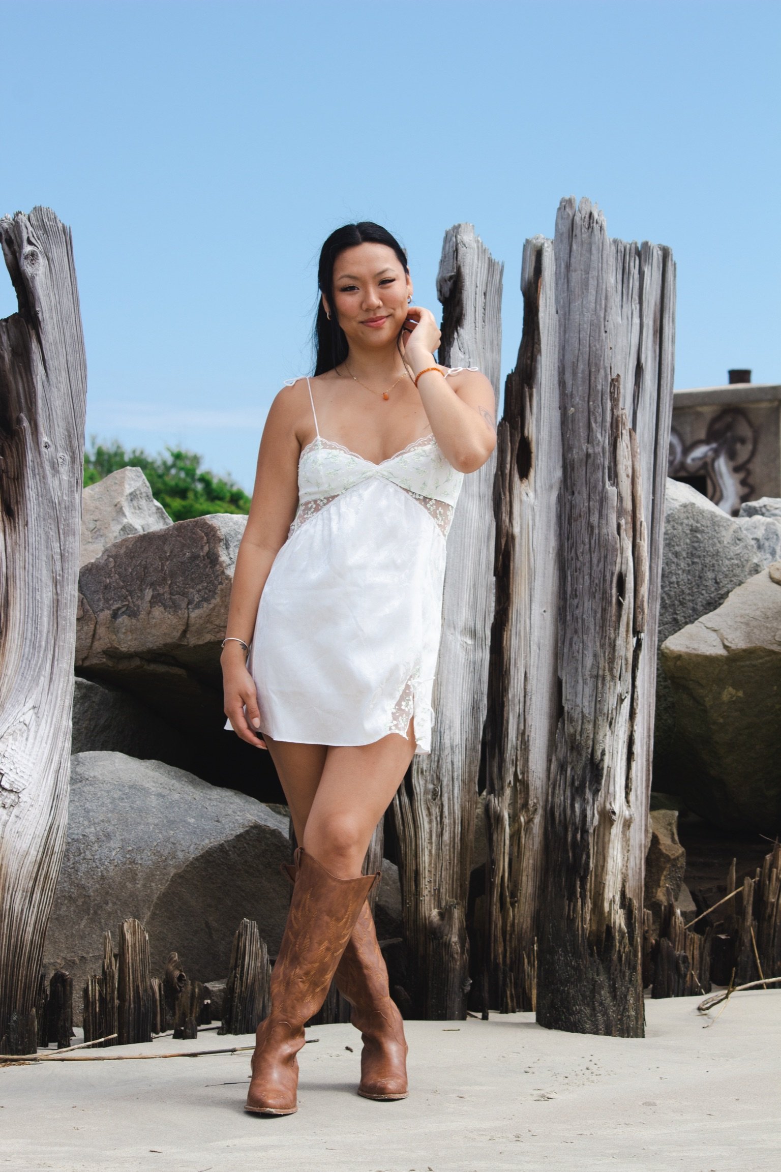 A graduate in a white dress standing on a sandy beach, wearing brown cowboy boots, with weathered wooden posts and rocks in the background under a blue sky. Shot on Sullivans Island, South Carolina.