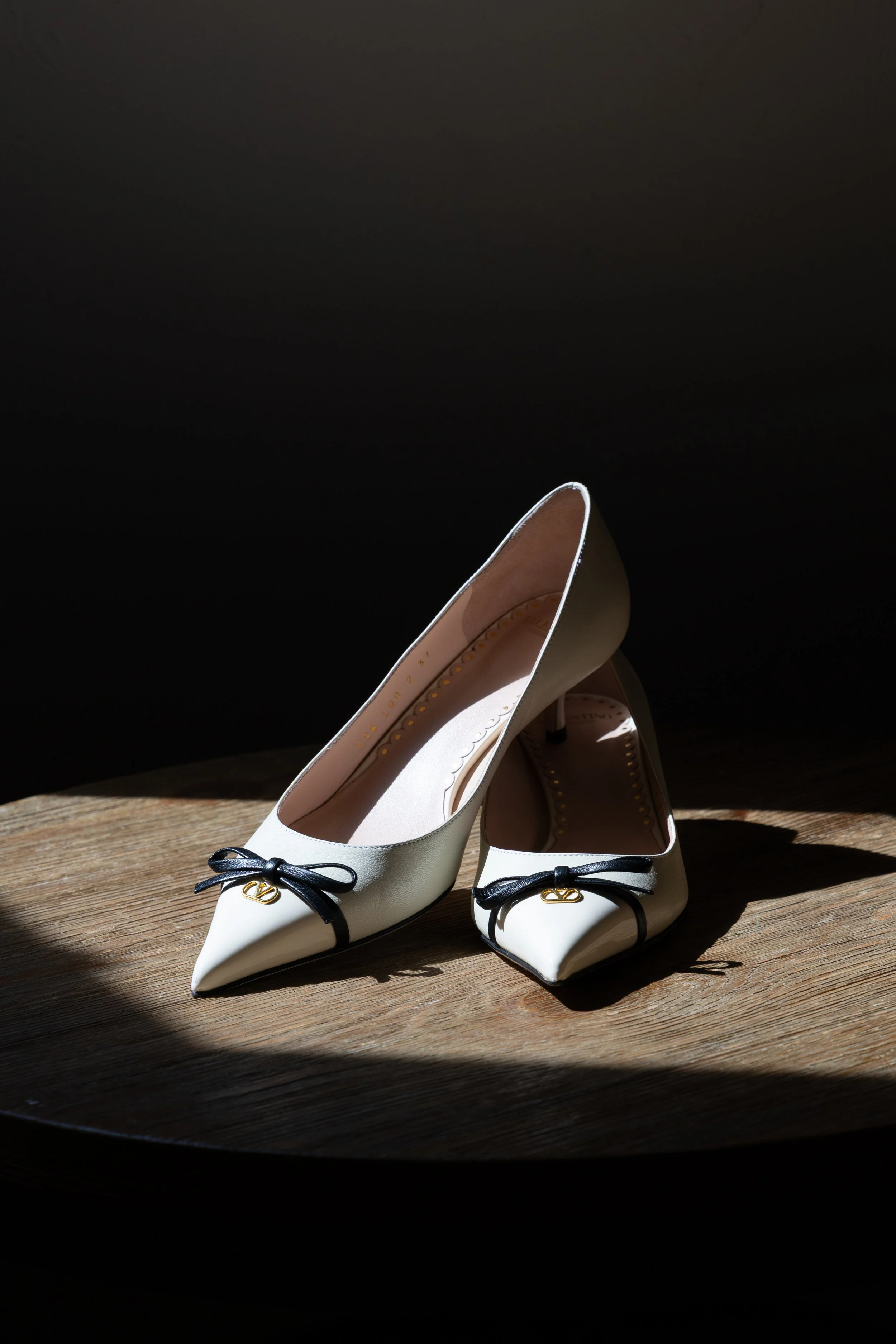 Pair of white high-heeled shoes with black bows and gold accents, positioned on a wooden surface with a dark background and light shining on them.