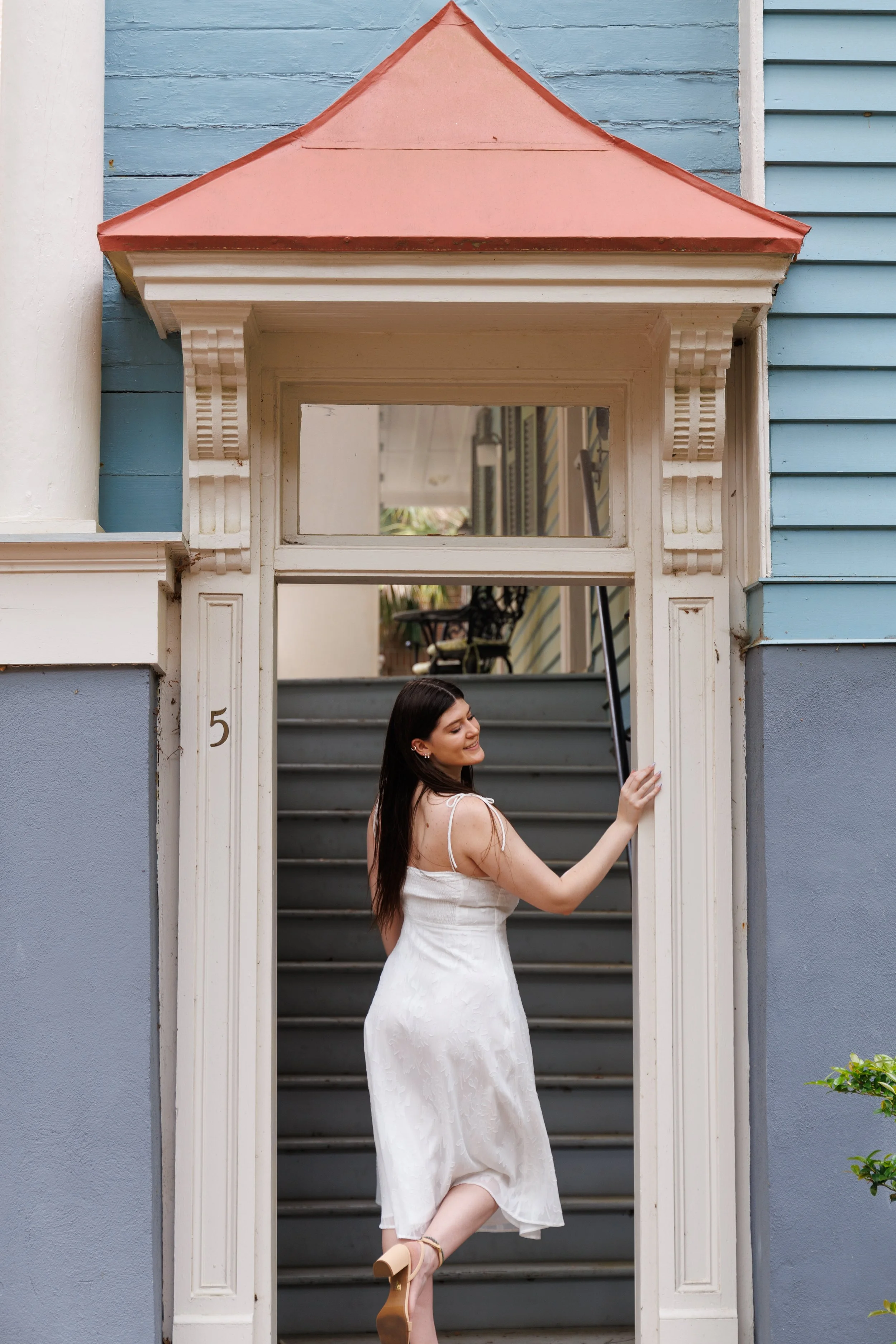 A woman wearing a white dress and beige heels, standing on a staircase, smiling and leaning against a doorframe of a house painted in pastel blue with a pink roof above the door.