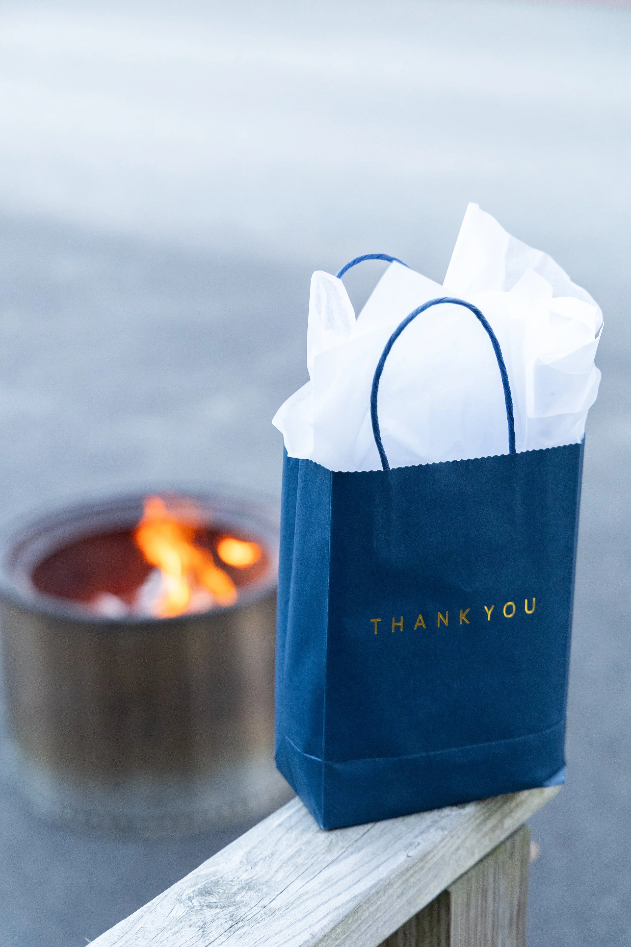 A navy blue gift bag with gold lettering saying 'Thank You' sitting on a wooden railing, with white tissue paper inside. A small fire is burning in a metal container in the background.