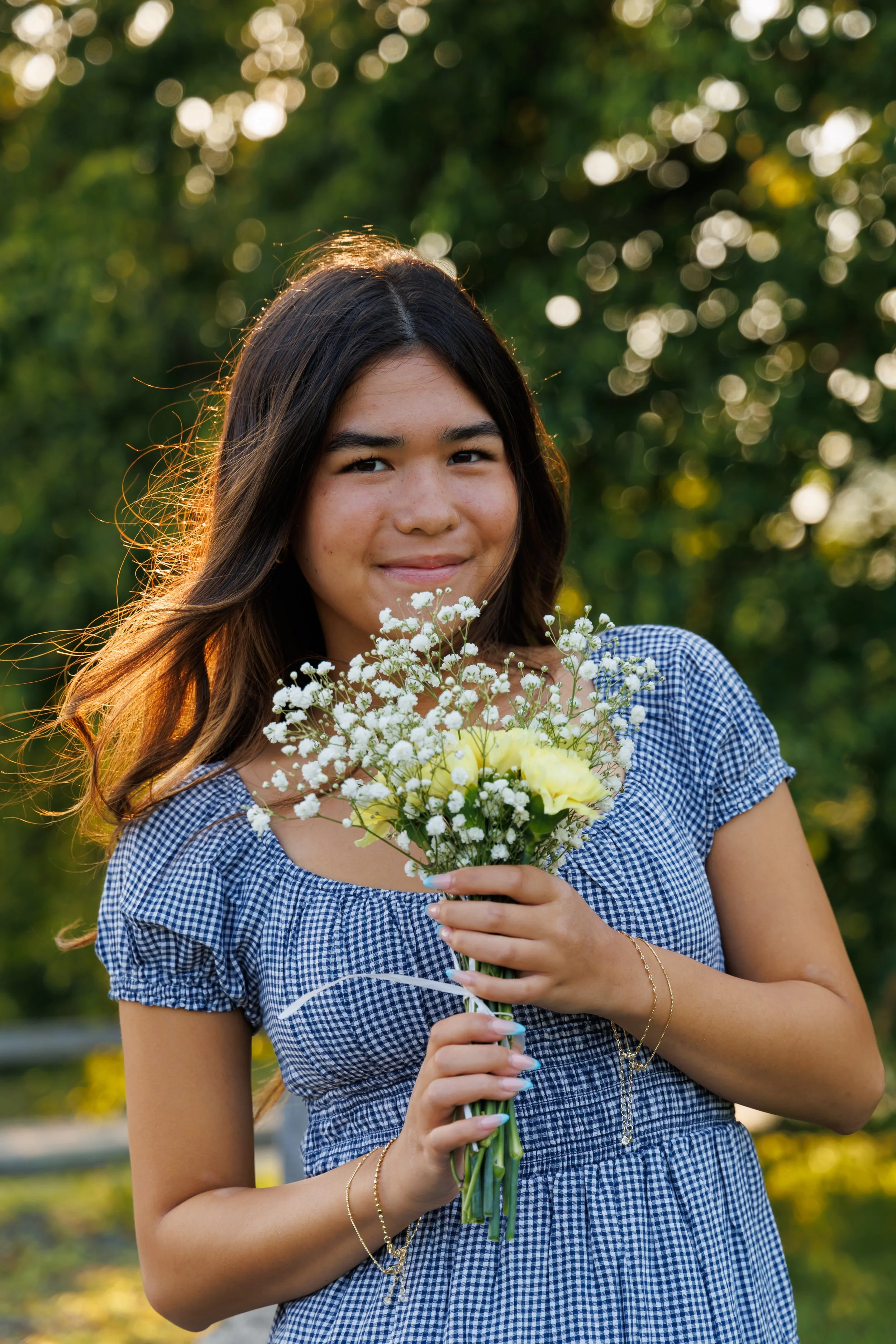 Young woman with long brown hair holding a bouquet of white and yellow flowers, outdoors during golden hour, wearing a blue checkered dress, smiling.