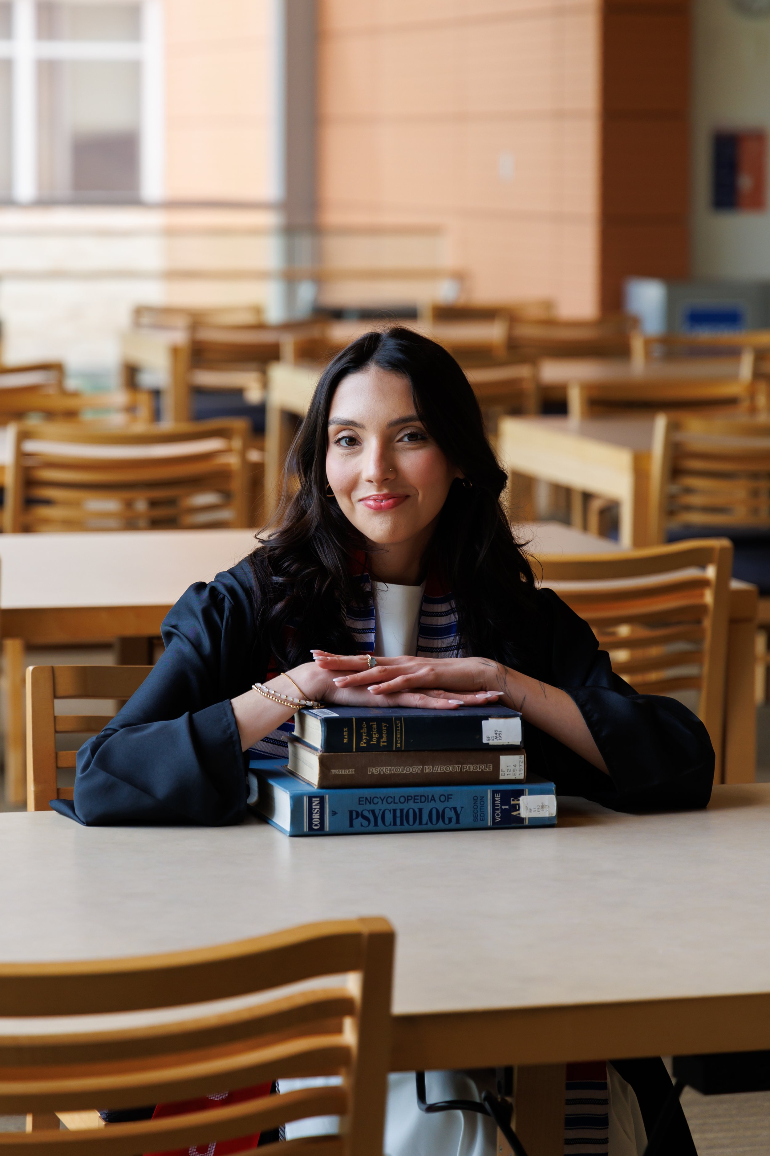 A young woman with dark hair and a nose piercing smiling at the camera, sitting at a table in a room with yellow and brown wooden chairs and tables, with a stack of psychology books in front of her.