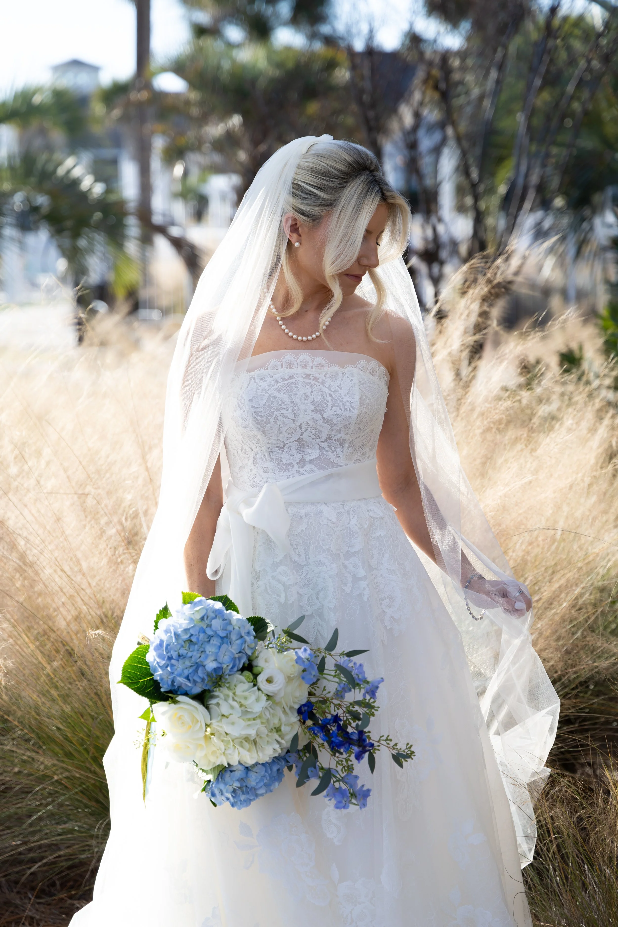 A bride in a white lace wedding gown with a pearl necklace and a sheer veil stands outdoors, holding a bouquet of blue and white flowers, with a background of trees and dry grass.