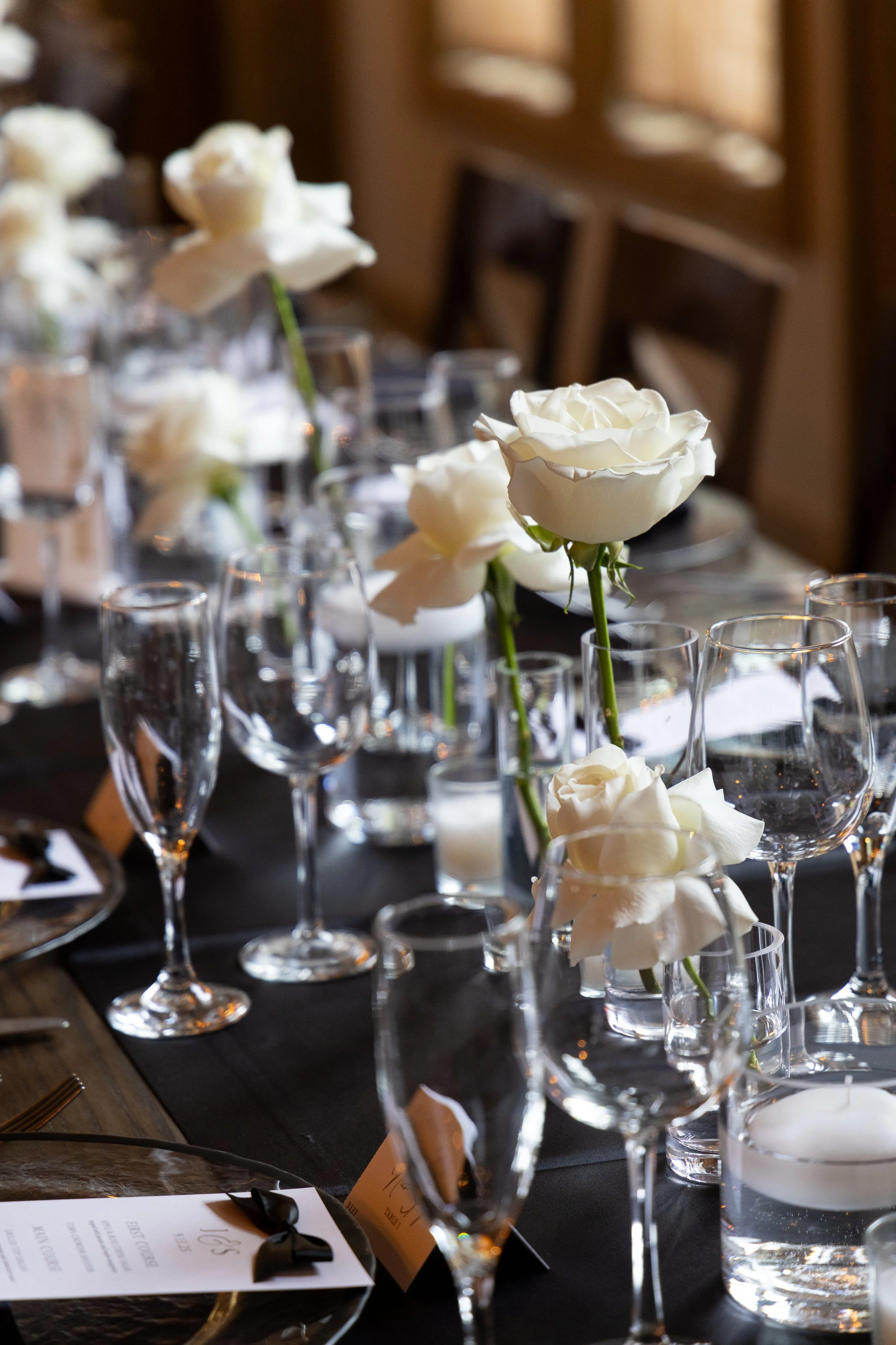 A table set with multiple clear wine glasses and white roses in small vases at a formal event or wedding reception.