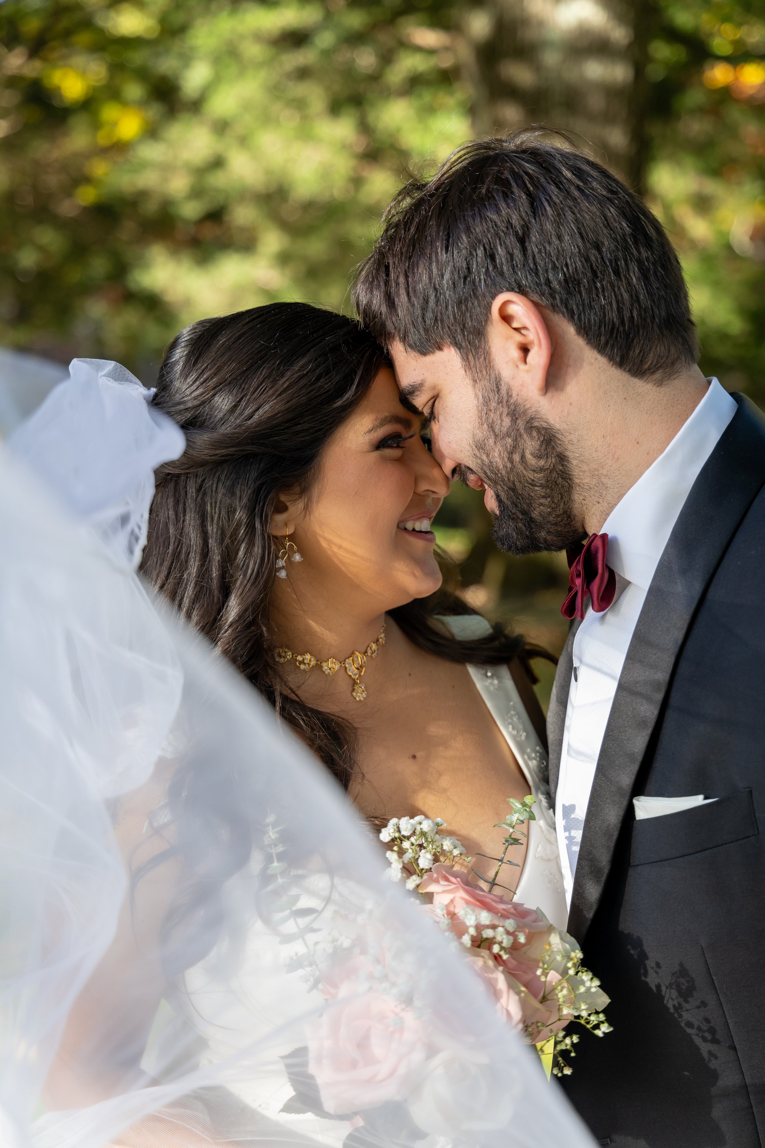 A bride and groom share a close moment during their wedding, touching foreheads and smiling in a joyful outdoor setting surrounded by greenery. Documentary style in the Greater Boston Area.