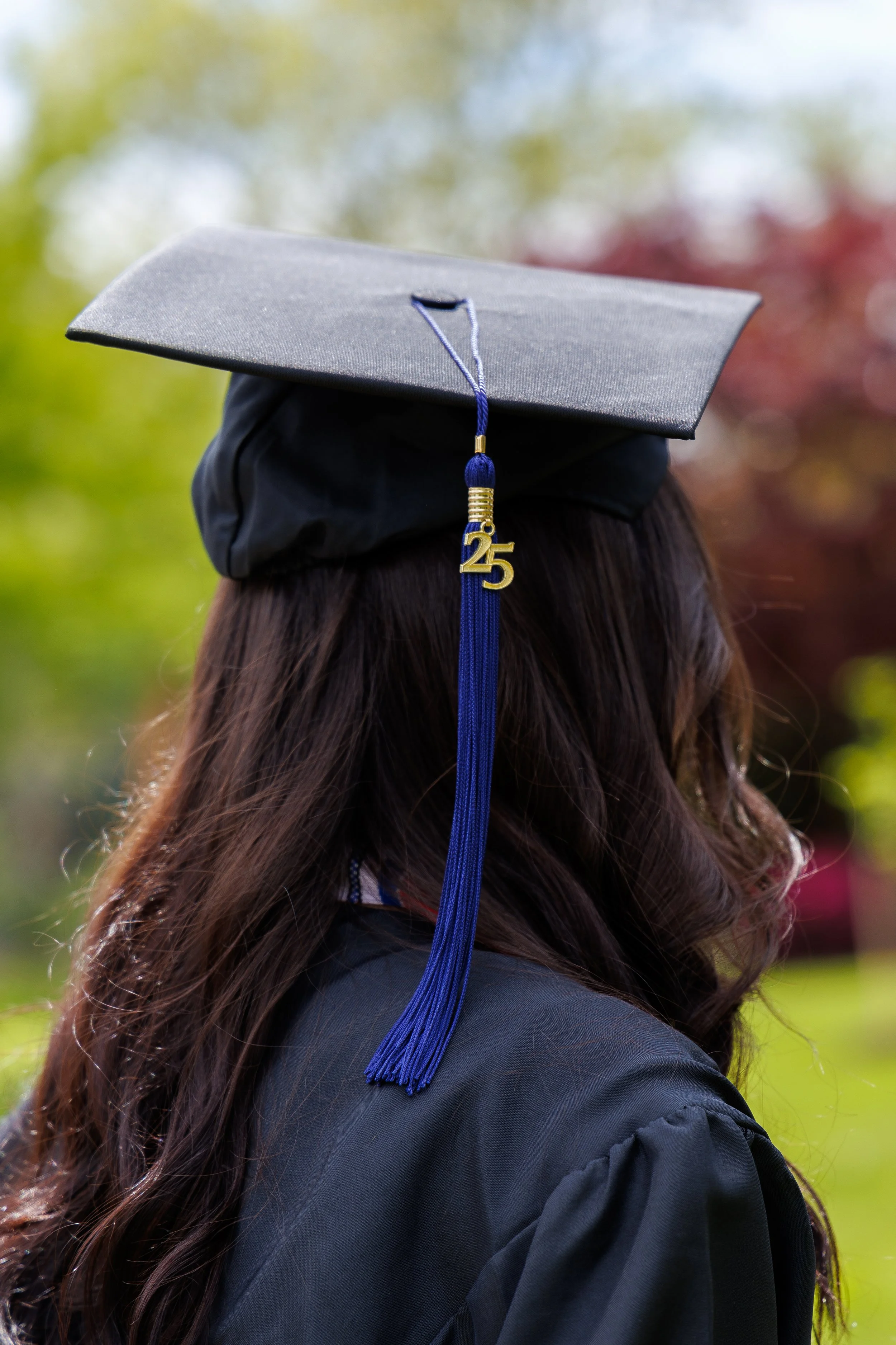 Side view of a person wearing a black graduation cap with a blue tassel and a gold charm that says '25', standing outdoors with blurred colorful trees in the background.