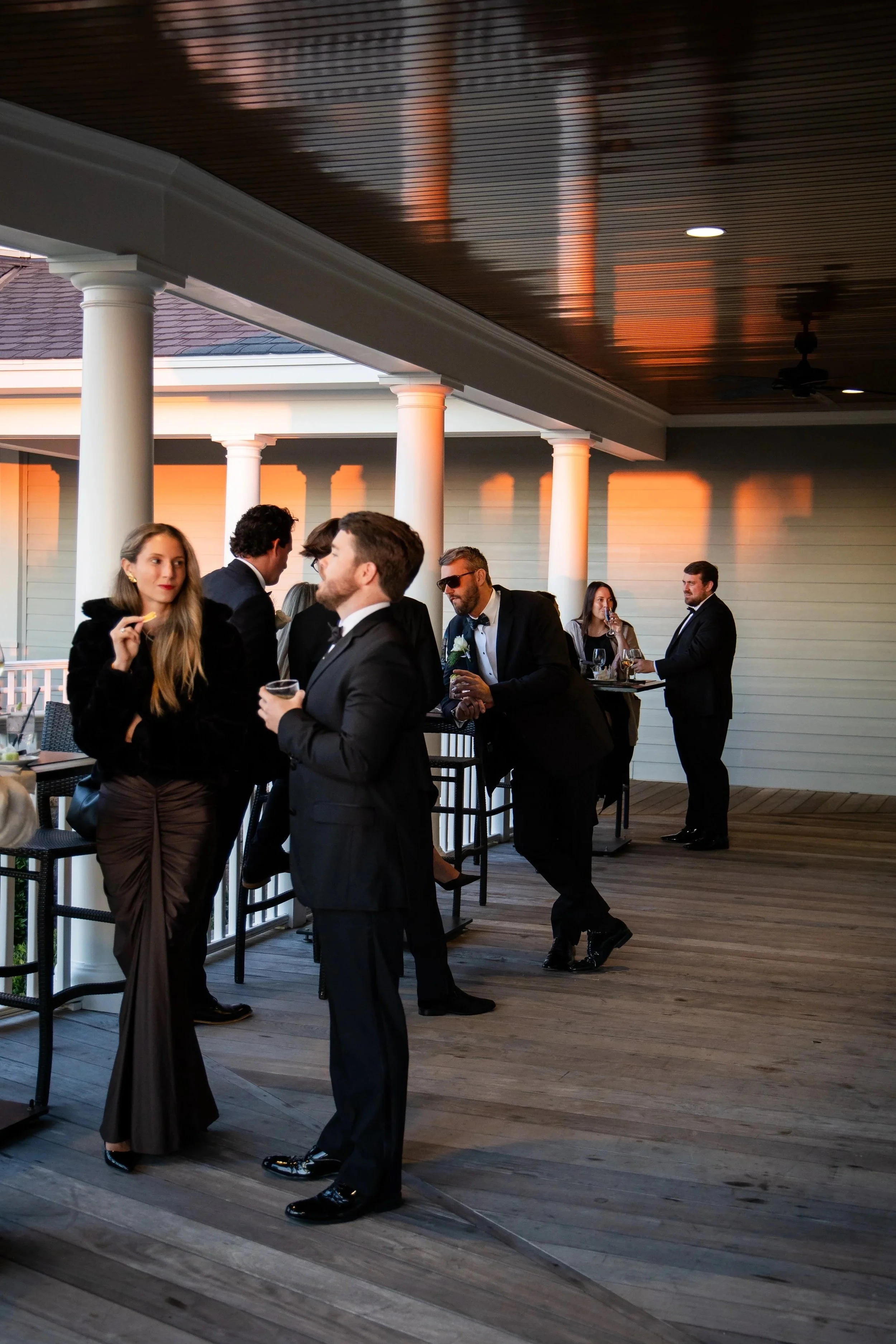 People dressed in formal attire socializing on a covered outdoor patio during sunset, with a wooden ceiling and white columns. Shot in Charleston, South Carolina.