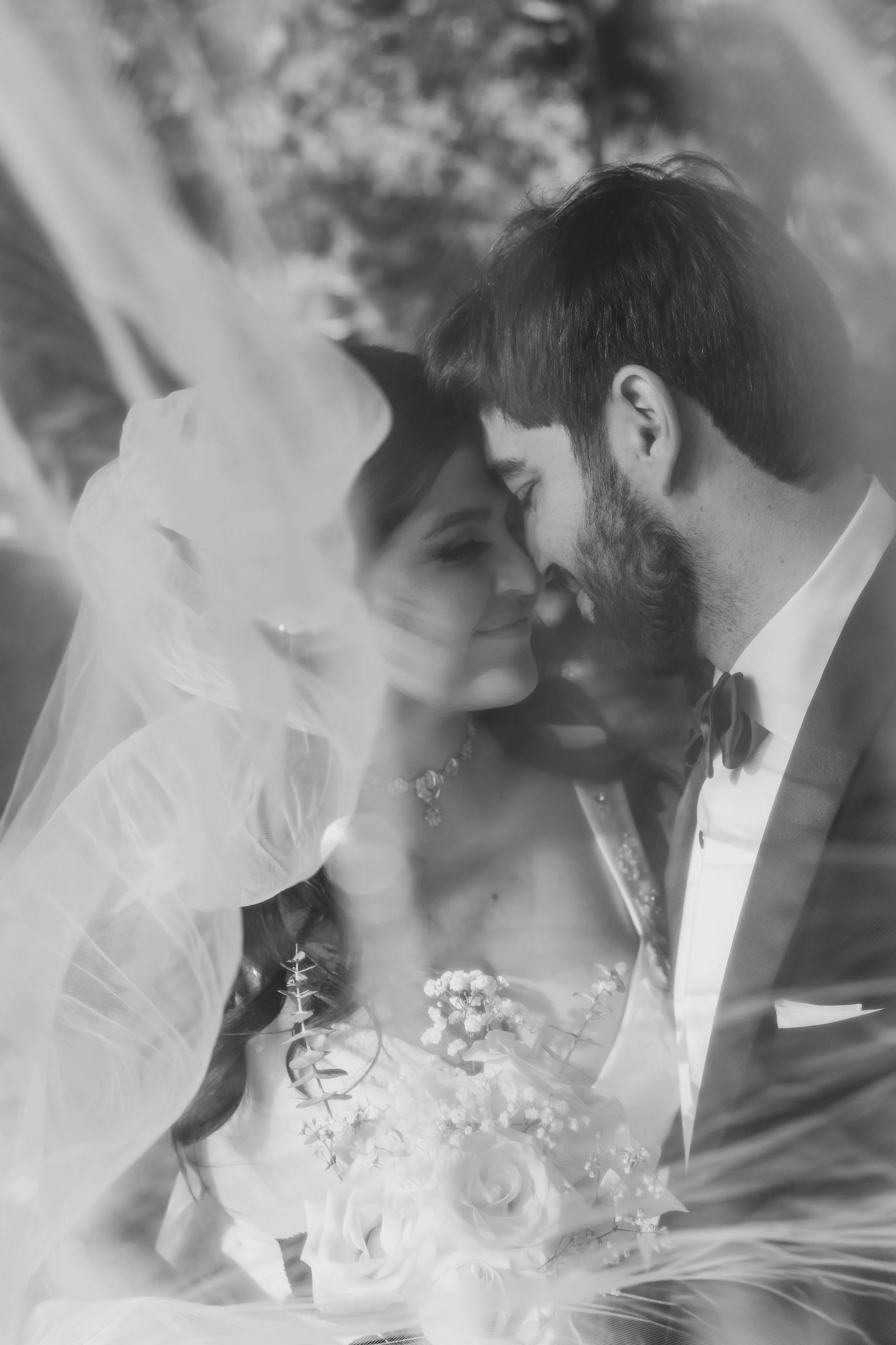 A black and white photograph of a bride and groom touching foreheads, smiling with eyes closed, with wedding flowers in the foreground. Shot in Boston, Massachusetts.
