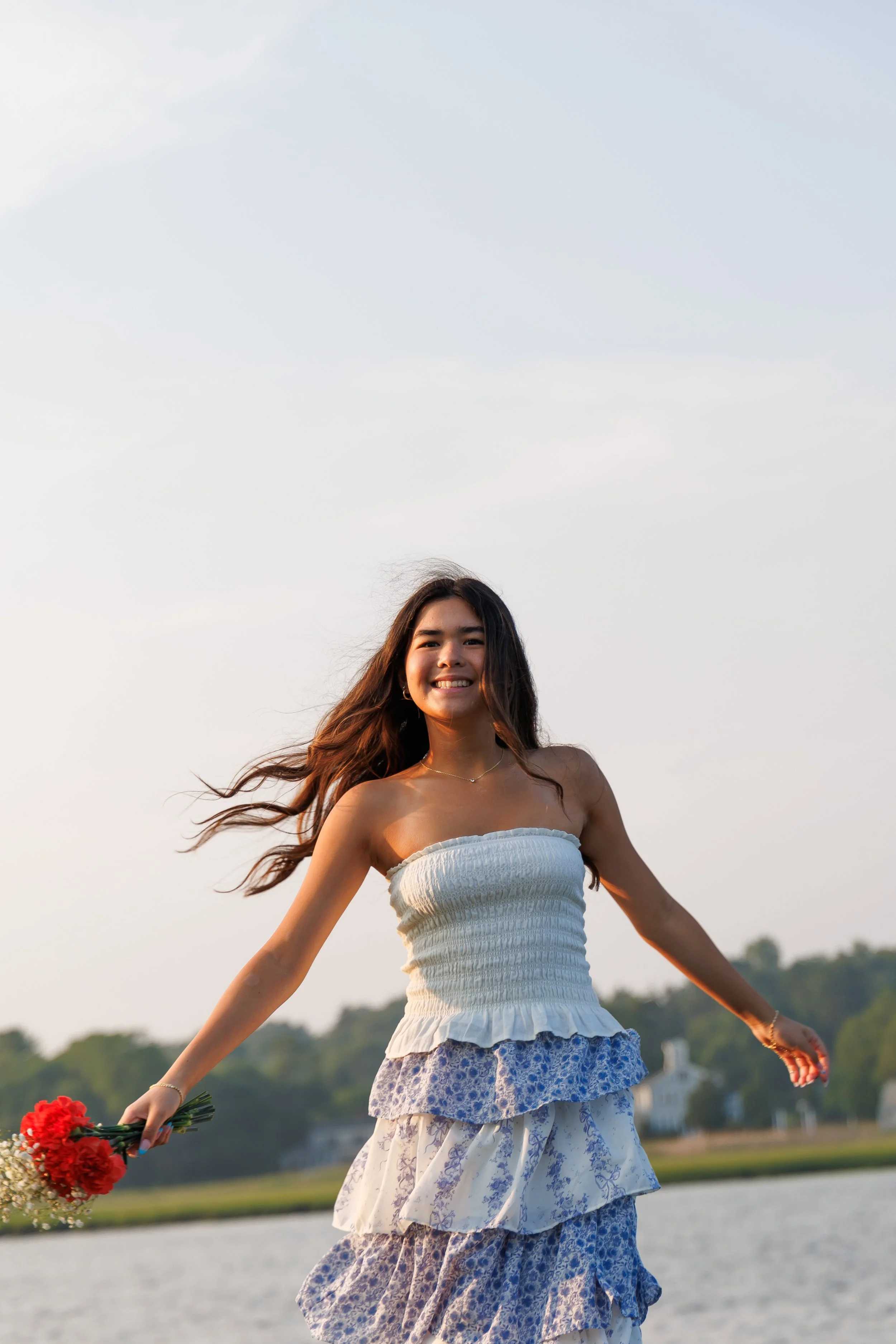 A young woman smiling and waving outdoors during sunset, holding a bouquet of red flowers, wearing a strapless white top and a layered blue floral skirt, with a body of water and trees in the background.