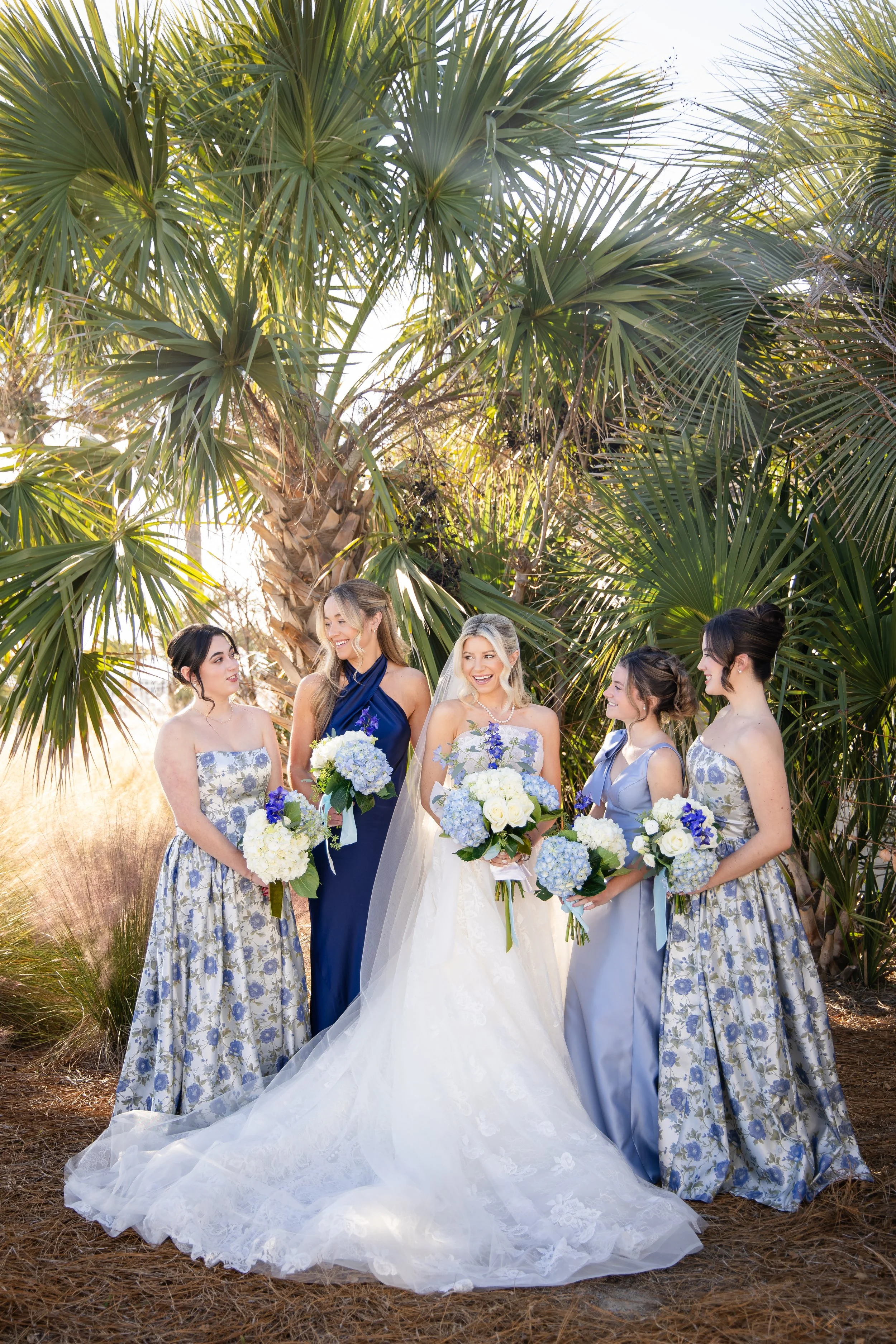 A group of women dressed for a wedding standing outdoors among large palm trees, smiling and holding white and blue floral bouquets. Shot at Seabrook Island, South Carolina.