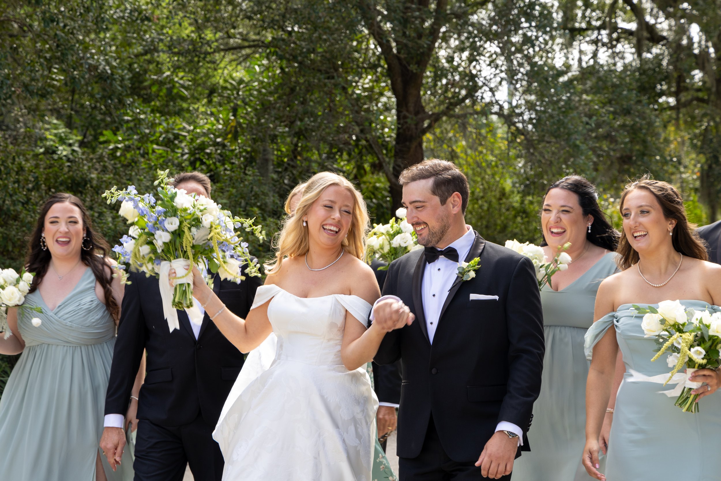 A bride and groom smiling and holding hands during their wedding ceremony, surrounded by bridesmaids in pastel dresses holding bouquets of white and purple flowers, in a lush outdoor setting. Shot at Hampton Park in Charleston, South Carolina.