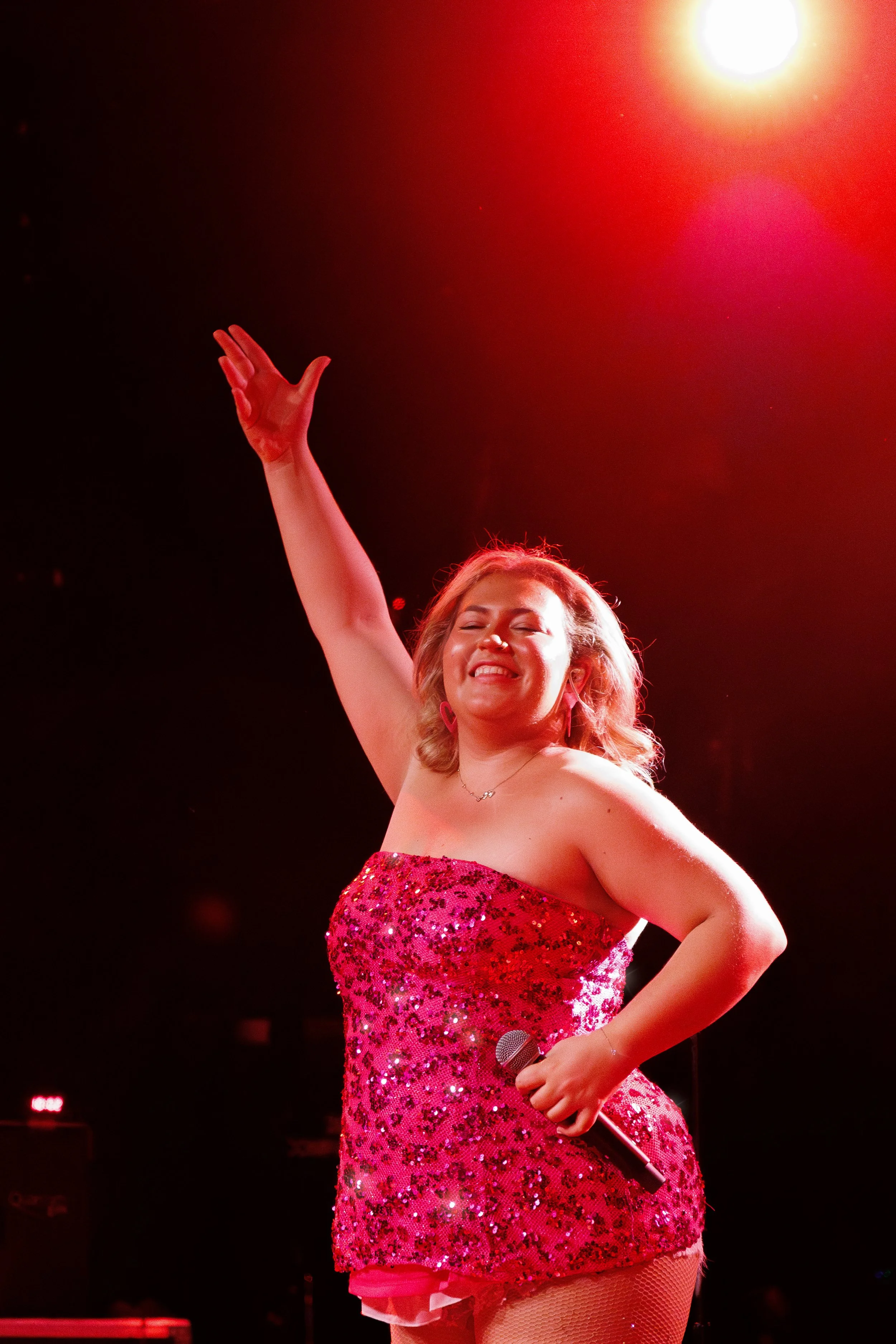 A woman on stage with a big smile, wearing a sparkling pink dress, holding a microphone, with her other arm raised, under a bright stage light.