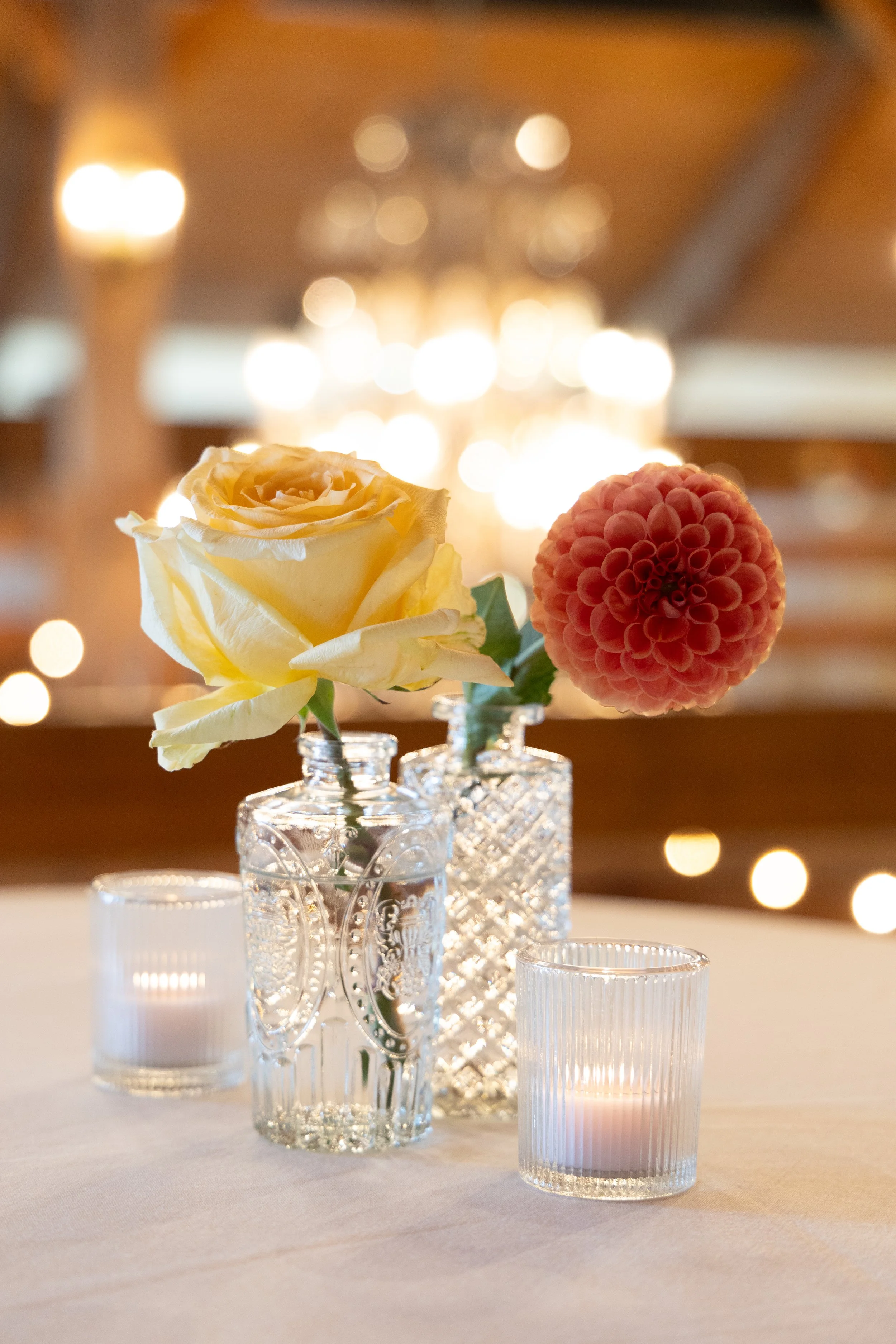 Close-up of a floral centerpiece featuring a yellow rose and pink dahlia in cut glass vases, surrounded by small lit candles on a white tablecloth, with warm blurred lights in the background.