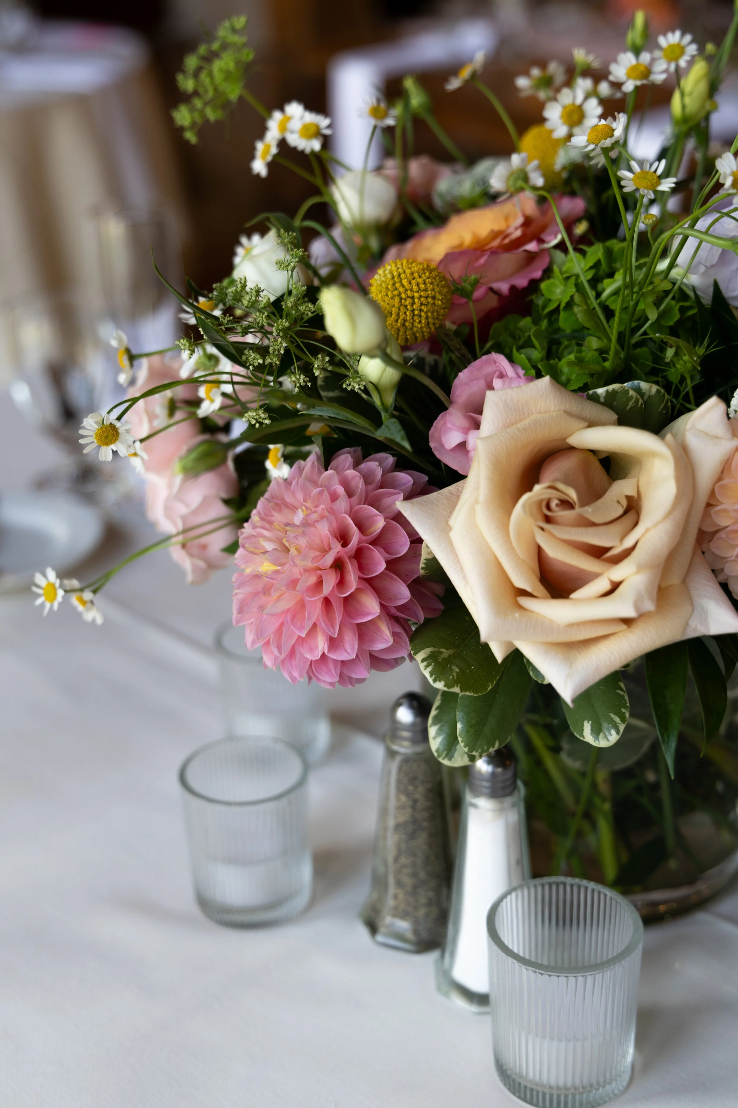 A floral arrangement at a wedding with white daisy-like flowers, pink and cream roses, yellow billy balls, and greenery in a glass vase, with empty clear glasses on a white table.