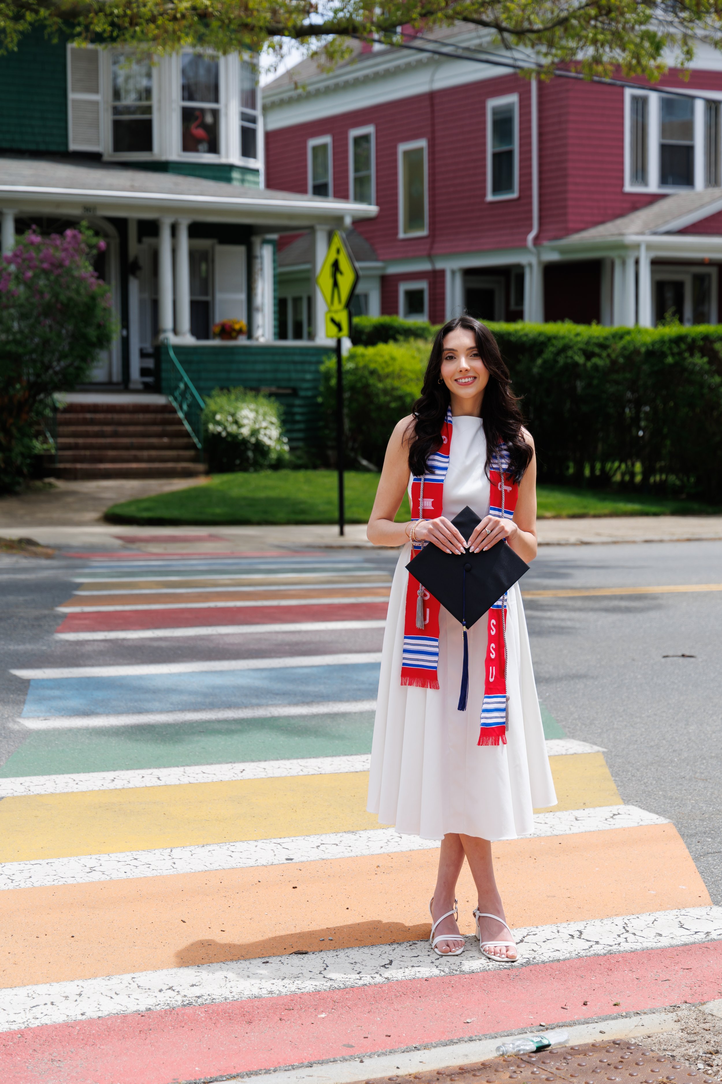 Young woman in a white dress holding a graduation cap, smiling, standing on a colorful rainbow crosswalk in front of colorful houses, with rainbow colors painted on the street.