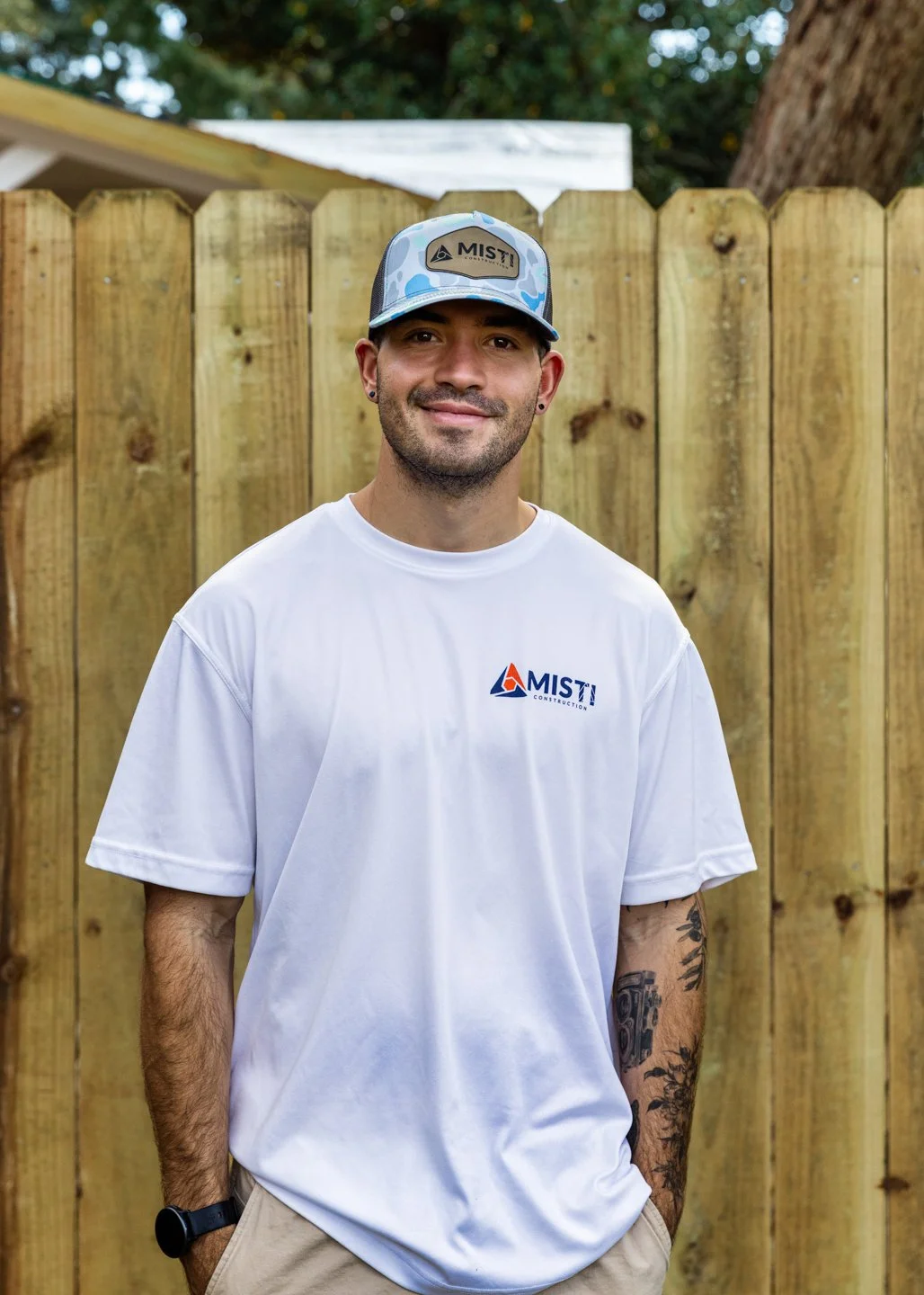 A young man with a beard and earrings, wearing a white T-shirt and a camouflage baseball cap, stands in front of a wooden fence. He has tattoos on his left arm and is smiling.