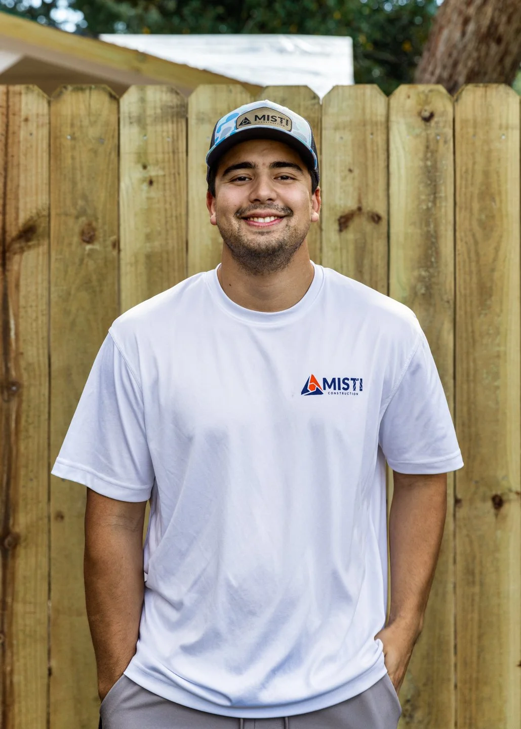 A young man wearing a white T-shirt and a blue and white cap, smiling with his hands in his pockets, standing in front of a wooden fence with a tree and house in the background.