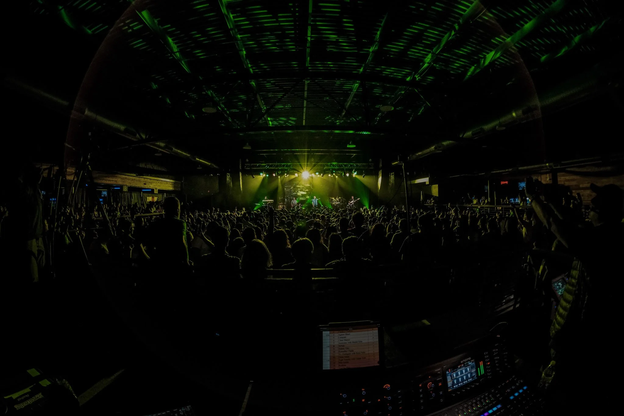 Audience watching a live concert with a band on stage illuminated by green and yellow lights, inside a large indoor venue.