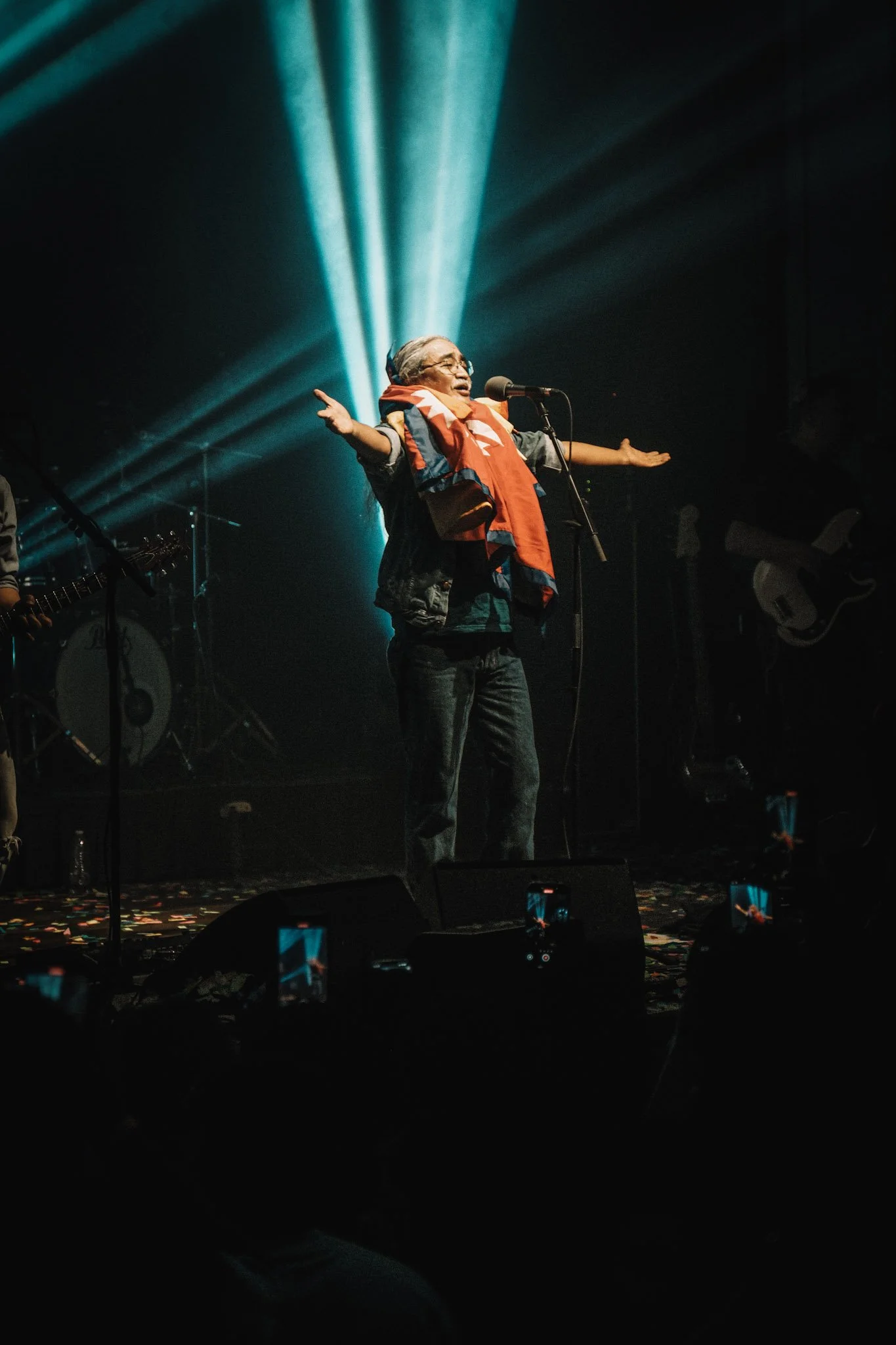 A person with silver hair and glasses singing into a microphone on stage, with arms outstretched, surrounded by stage lights and musical instruments in the background.