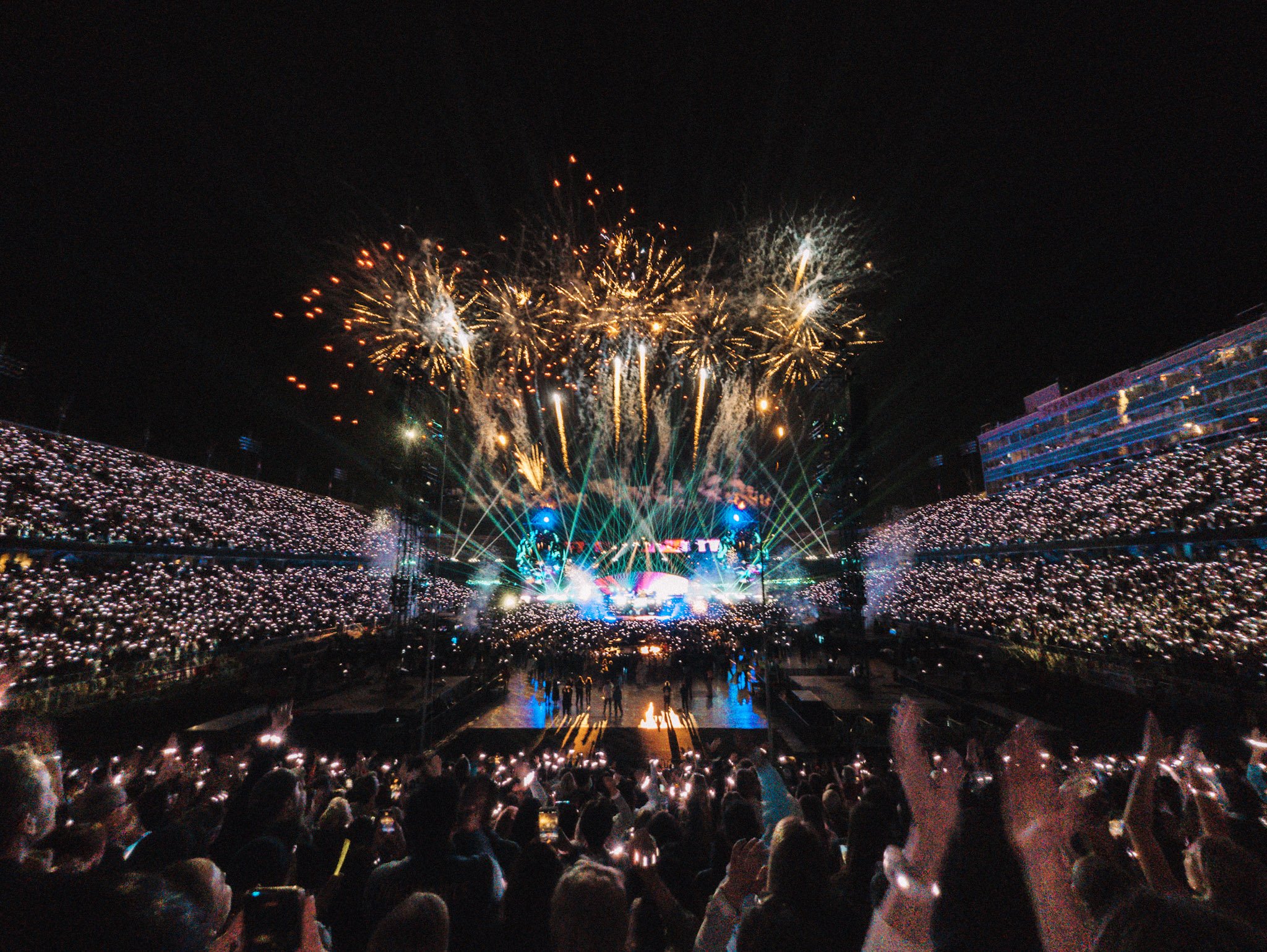 Crowd at a concert or festival night event with fireworks display overhead, many people holding up lights or phones.