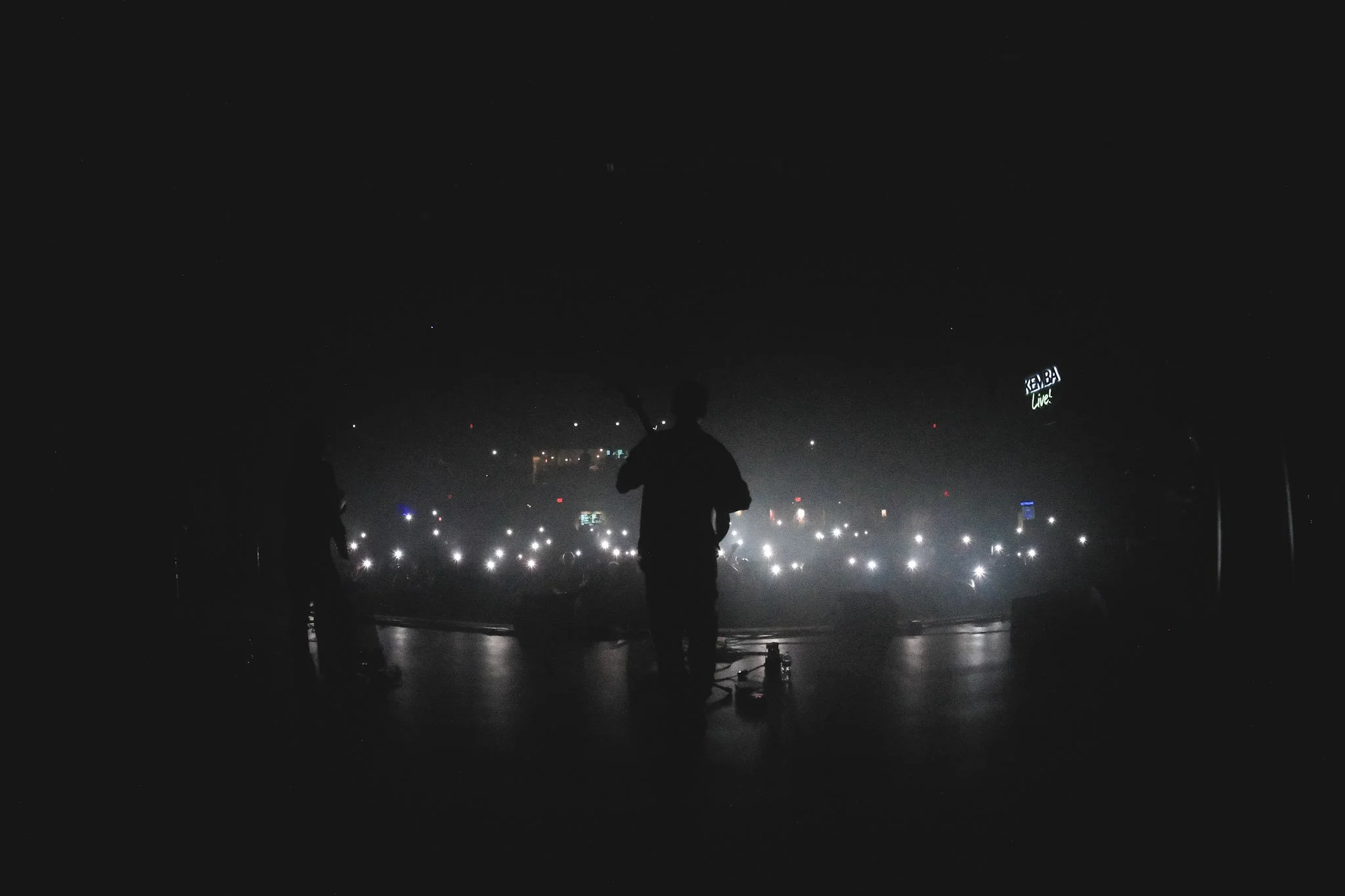 Silhouette of a performer on stage with a microphone in hand, facing an audience with lights and screens in the background.