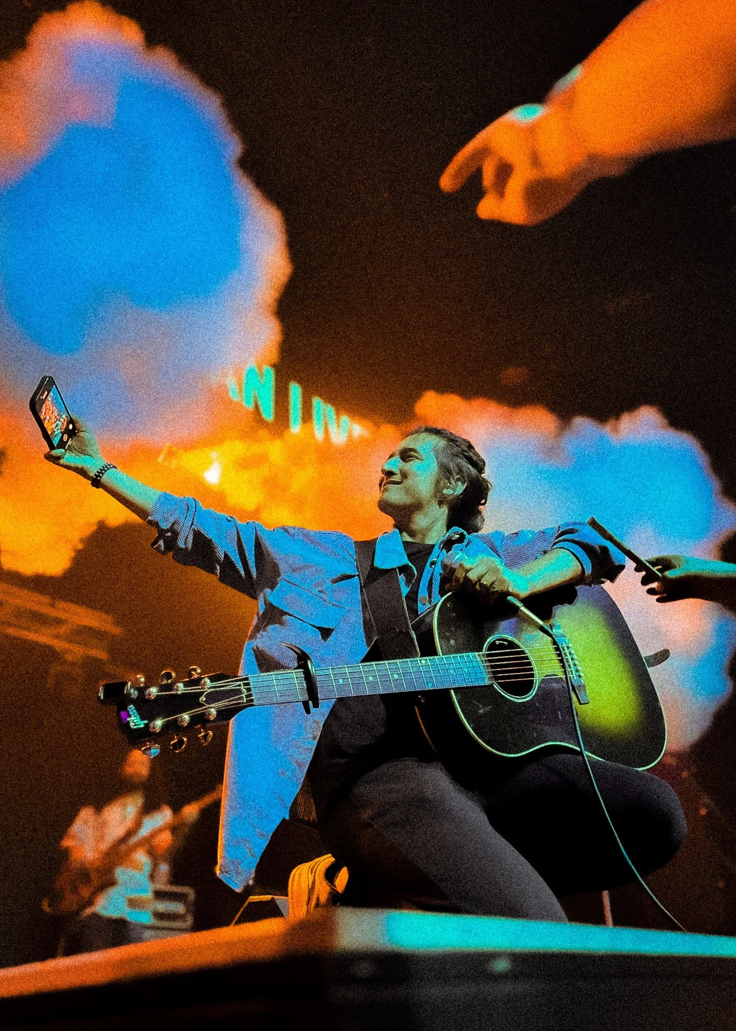 A young man with long hair playing an acoustic guitar on stage, smiling, with colorful stage lights and clouds of smoke behind him, and another person taking a selfie with a smartphone.