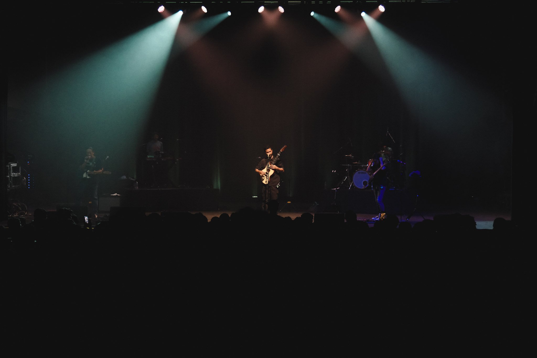 A live concert scene with a band performing on stage illuminated by colored spotlights, with the audience visible in the foreground.